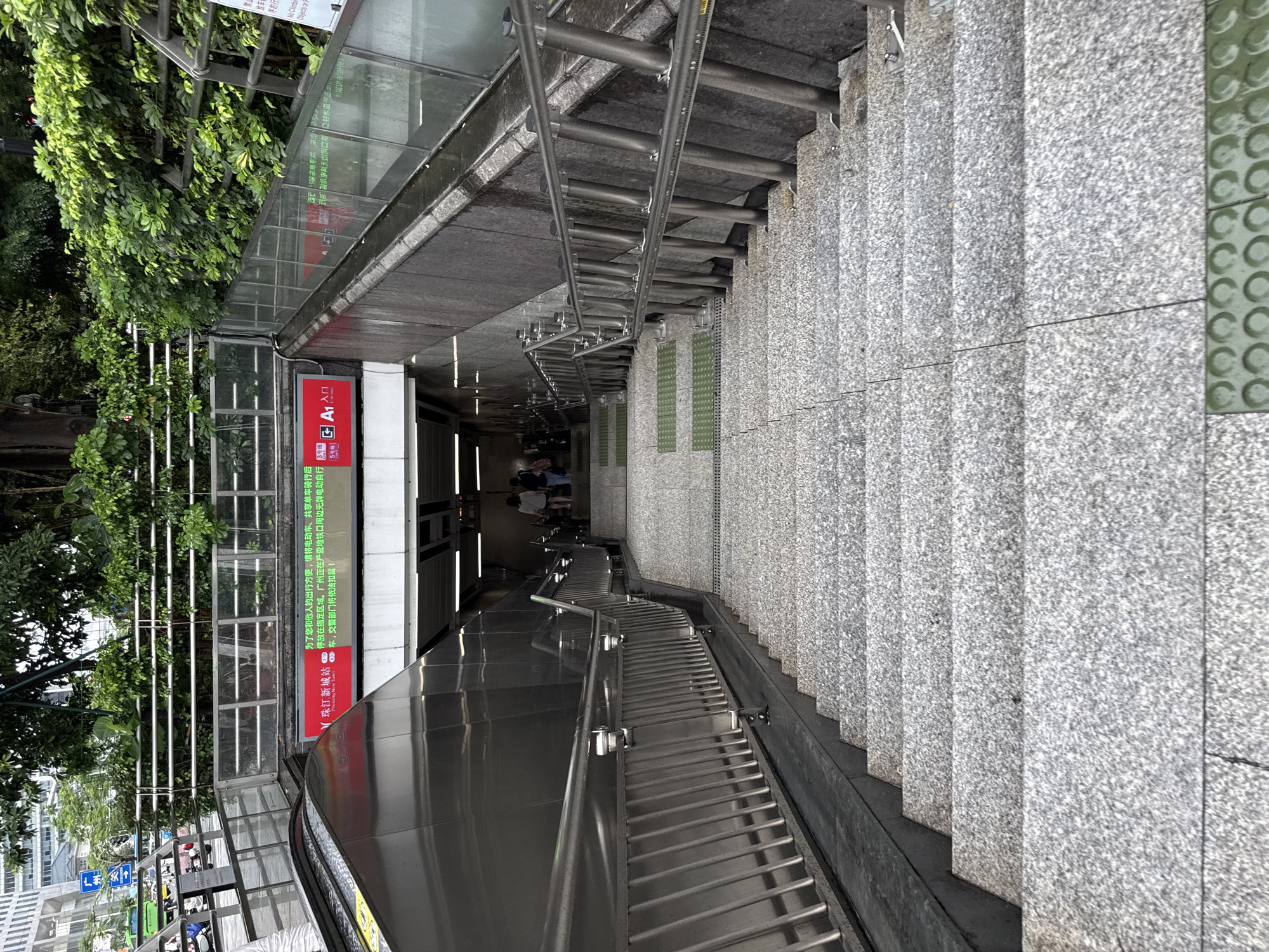 The image shows a downward view of a wide, outdoor concrete staircase in Tianhe District, China. The staircase is flanked by metal railings and concrete walls. At the top of the stairs, an escalator is visible to the left, and a glass barrier separates the escalator from the stairs. Above the entrance to the stairs, a red digital display board shows Chinese text and logos, indicating the Zhujiang New Town Station and the A1 entrance. Green plants and trees are visible at the top of the scene, with buildings in the background. Two individuals are seen descending the stairs in the distance. The environment appears to be a public transportation hub.