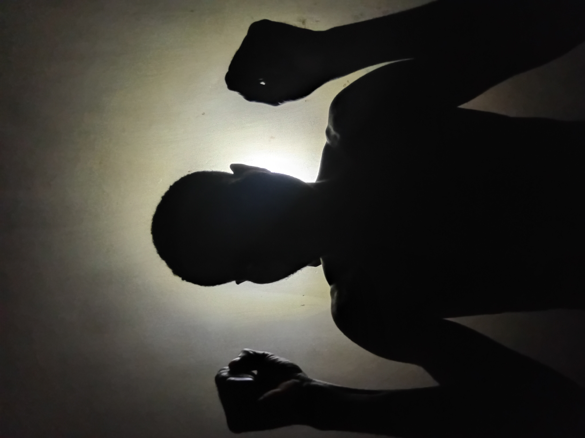 A low-angle shot in Ikorodu, Nigeria, captures a silhouette of a shirtless person with clenched fists raised. A light source behind the person creates a halo effect.