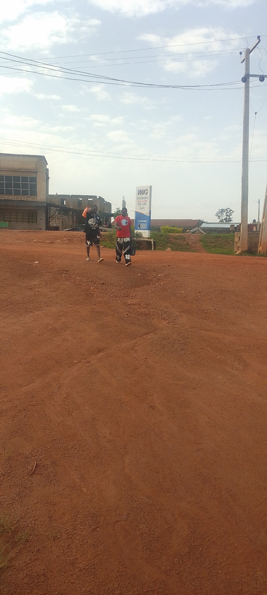 The image depicts an outdoor scene on a dirt road in Jos, Nigeria. Two men are walking away from the camera. The man on the left is wearing a black t-shirt with white lettering and black shorts, and he is carrying an orange object over his shoulder. The man on the right is wearing a red t-shirt and black pants with white stripes, and he is carrying a dark bag in his right hand. Behind them, a large sign with the text "NOC NGOBANG OIL & GAS NTG. LTD. Dura ACCESSORIES PLANT" is visible. To the left of the sign is a building with a facade of windows and corrugated metal roofing. Further in the background, more buildings and vegetation are present. A utility pole with electrical wires stands to the right of the frame. The ground is a reddish-brown dirt, characteristic of the soil in some parts of Jos. The sky is partly cloudy.