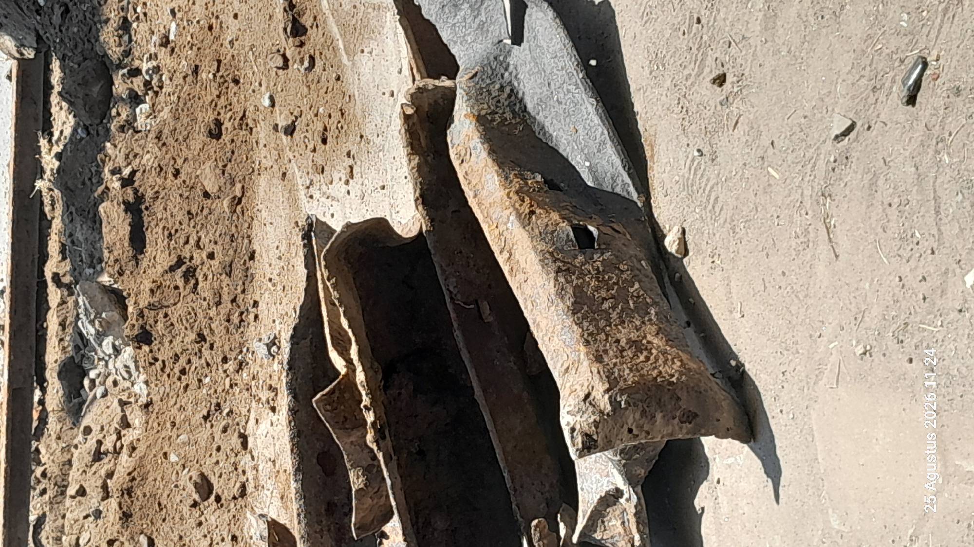 The image depicts a close-up, eye-level view of several rusted metal beams and debris on a sandy, concrete-like surface. The beams are of various sizes and shapes, some appearing to be structural components, with significant oxidation and wear. In the background, piles of dirt and smaller rocks are visible, suggesting a construction or demolition site. The lighting indicates it is daytime, casting shadows on the ground. A timestamp in the lower left corner reads "25 Agustus 2026 11.24". The location is identified as Jomblang, Indonesia.