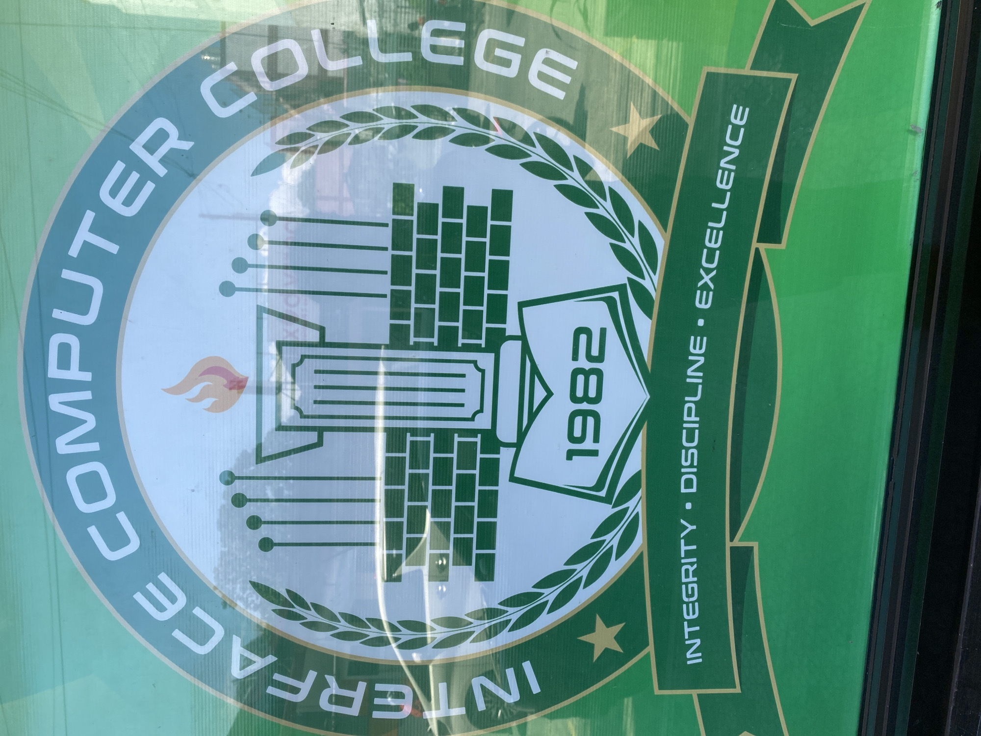 The image displays a prominent circular logo for an educational institution, primarily in shades of green and white, set against a bright green background. The logo appears on a sign or banner, with visible reflections indicating it might be behind glass or a translucent material.
The outer ring of the circular logo features white uppercase text that reads "COMPUTER COLLEGE" along the top curve and "INTERFACE" along the bottom curve, identifying the institution as "INTERFACE COMPUTER COLLEGE."
The central emblem of the logo is rich in detail. It includes a stylized building with columns, and to its right, a structure composed of stacked rectangular blocks, possibly representing data or bricks. A torch with an orange-red flame is positioned on the left side of this central design. Circuitry-like lines and small dots extend from these elements. On the right, a book is depicted with the year "1982" written vertically on its cover. This entire inner design is framed by a green laurel wreath. Two yellow stars are placed symmetrically outside the wreath, one on each side.
To the right of the main circular logo, a dark green banner or ribbon extends diagonally downwards. This banner features white text that reads "INTEGRITY • DISCIPLINE • EXCELLENCE," with dots separating the words. The overall impression is that of an institutional emblem emphasizing technology, history, and core values. No people or activities are visible in the photograph; it solely captures the static sign.