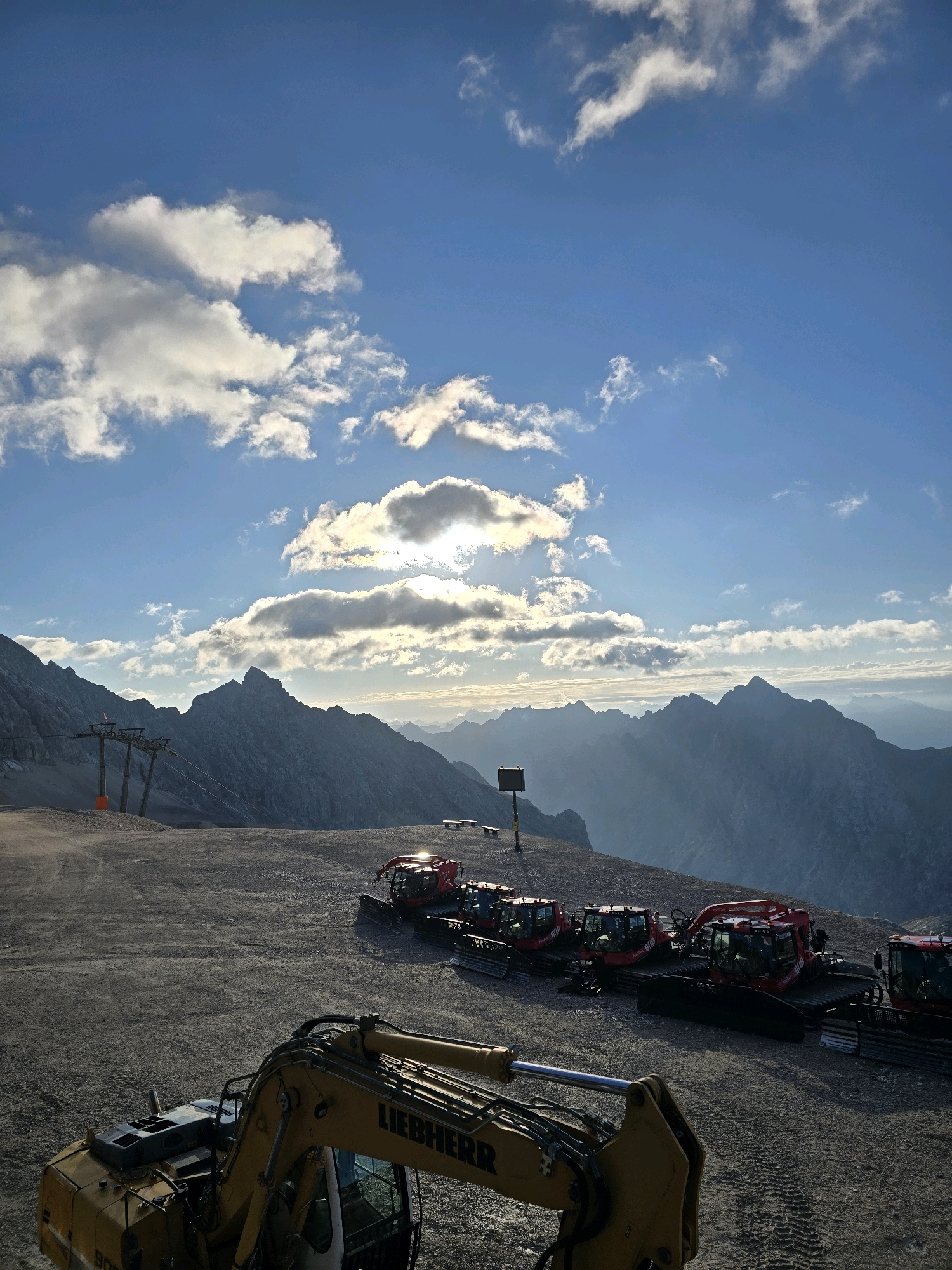 This image captures a daytime scene at the Zugspitze, Germany's highest peak, located near Garmisch-Partenkirchen. In the foreground, a yellow Liebherr excavator is parked on a gravel surface. Behind it, a line of red snow groomers is visible, parked parallel to each other. The background is dominated by a majestic mountain range, with the Zugspitze prominently featured. The sky is a vibrant blue, with scattered white clouds, some of which are illuminated by the sun, creating a dramatic lighting effect. A ski lift structure with cables is visible on the left side of the image. The overall impression is one of an alpine landscape, possibly during a period of maintenance or preparation for winter activities, given the presence of construction and snow-grooming equipment. There are no people visible in the image.