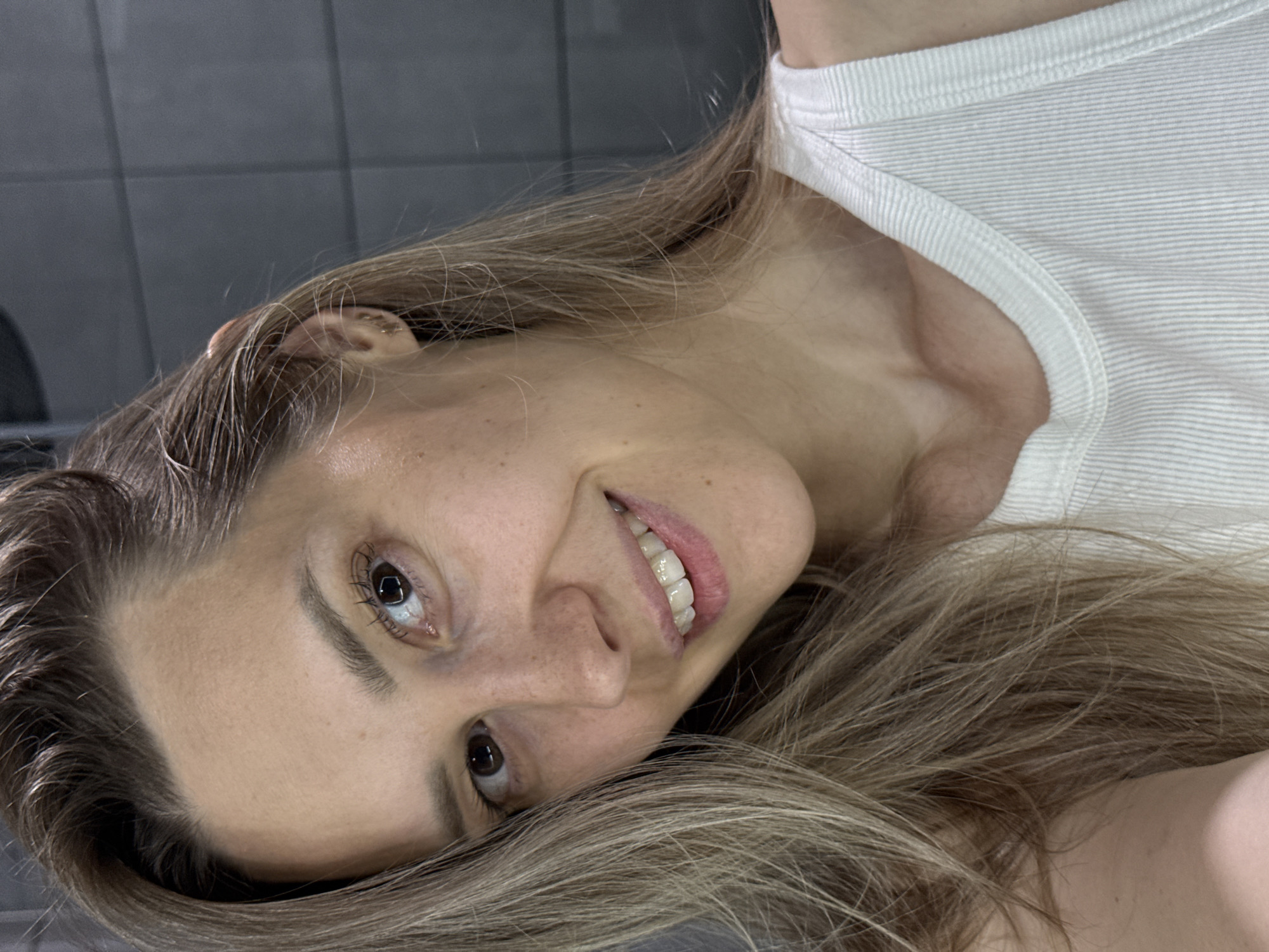 A woman with long, blonde hair is lying down, smiling at the camera. She is wearing a white shirt. The background is a gray tiled wall. The location cannot be determined from the image.