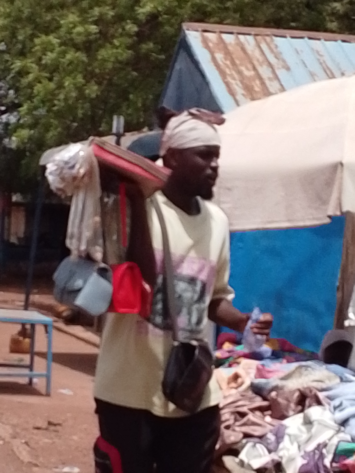 In a sunlit outdoor setting in Tamale, Ghana, a dark-skinned man with a headscarf is seen carrying goods. A variety of bags hang from his shoulder. In his right hand, he holds a small plastic item. Adjacent to him is a table laden with folded fabrics. The backdrop features a building with a blue and rusted roof and a white umbrella. Trees are visible in the distance.