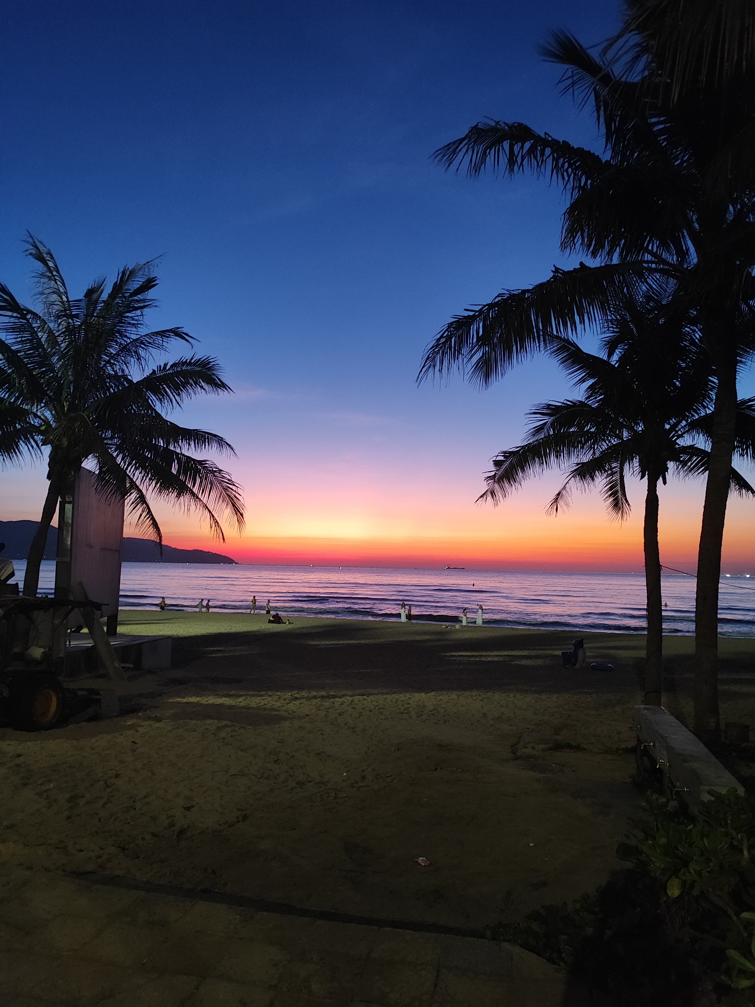 The image depicts a beach scene at dusk in Đà Nẵng, Vietnam. The sky transitions from a deep blue at the top to vibrant hues of pink, orange, and yellow near the horizon, indicating the sun has recently set. Two silhouetted palm trees frame the view, their fronds casting shadows on the sandy beach. The ocean stretches out before the beach, with gentle waves lapping at the shore. In the distance, a few small boats are visible on the water, and the outline of mountains can be seen against the colorful sky. Several people are scattered across the beach, some walking or sitting, while others appear to be engaged in activities near the water. The foreground consists of sand with some scattered debris, and to the left, a piece of heavy machinery and a wooden structure are present. The overall atmosphere is serene and tranquil, capturing the beauty of a tropical sunset.