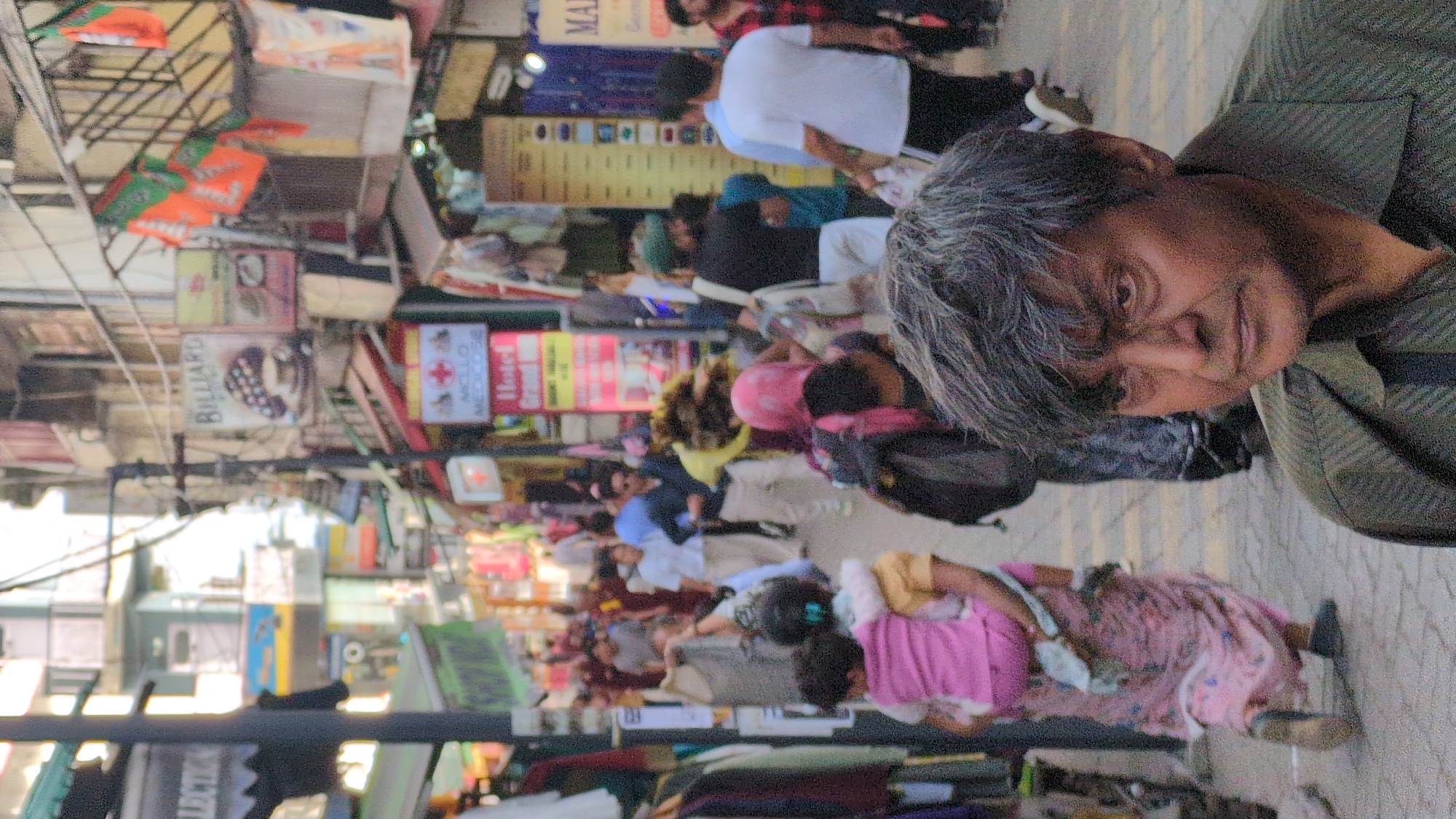 The image shows a crowded street scene in Dharamshala, India. Pedestrians of various ages and attires are walking along a narrow lane. In the foreground is an older person, presumably male, with gray hair and a muted green shirt, facing the camera. The street is lined with buildings and various shops indicated by colorful signs, including one for "Billiard" and a hotel. Overhead are power lines and strung flags. The ground is paved.