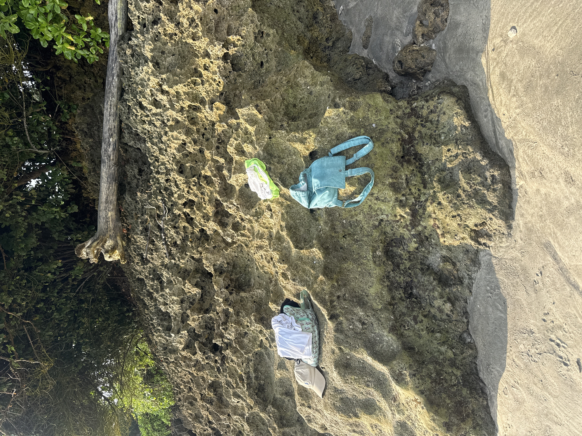 The scene is outdoors, likely near a beach in Jimbaran, Indonesia. A large, weathered rock formation dominates the view. The rock's surface is porous and uneven. To the left, vegetation including trees and shrubs are visible. Scattered on the rock are personal items: a blue handbag with handles, a pair of green and white shoes, a white t-shirt, a patterned shirt, and a tan baseball cap. A patch of sand is visible along the right edge of the rock formation.