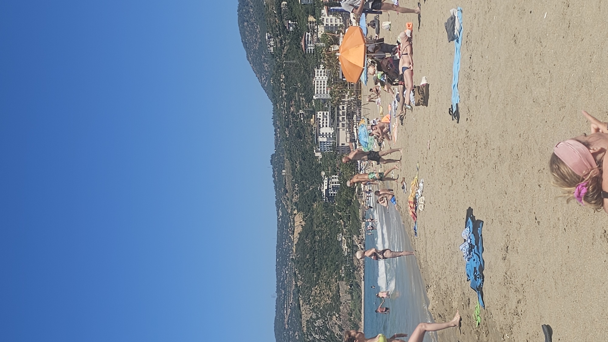 An overhead shot shows a crowded beach in Alanya, Turkey, under a clear blue sky. The sand is filled with people sunbathing on towels and in chairs. Some individuals are wading in the shallow water, while others are walking along the shore. An orange umbrella provides shade for a group of people. Buildings and lush green hills form a backdrop to the beach. The image captures a lively summer day with many visitors enjoying the coastal scenery.