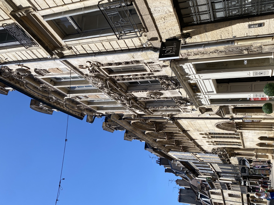 This image captures a street scene in the city of Dijon, France, specifically featuring the historic Logis du Roy building on the left, which houses the Tourist Office. The perspective is a low-angle view looking down a narrow, sunlit street lined with multi-story stone buildings under a clear, cloudless blue sky.
On the immediate right, a building with an ornate limestone facade dominates, featuring elaborate Baroque or Neoclassical carvings, decorative window surrounds, and closed and open shutters. A black rectangular sign displaying "joli caprice" in white text hangs from this structure. At ground level, a shop entrance is visible, flanked by two spherical green topiaries.
On the left, further down the street, stands a prominent stone building characterized by an upper-level arcade composed of a series of rounded arches, typical of Romanesque architecture. Beyond this, a continuous row of historic stone buildings extends into the distance, many featuring windows with blue or grey shutters, wrought-iron balconies, and rooftops with numerous chimneys and television antennae.
The street level shows a paved surface with several pedestrians. One older woman with white hair, wearing a light-colored top and carrying a light blue bag, walks towards the foreground on the left side of the street. Other indistinct figures are visible further in the background. A single black wire spans horizontally across the upper portion of the image. Bright daylight illuminates the scene, casting defined shadows on the right side of the street.