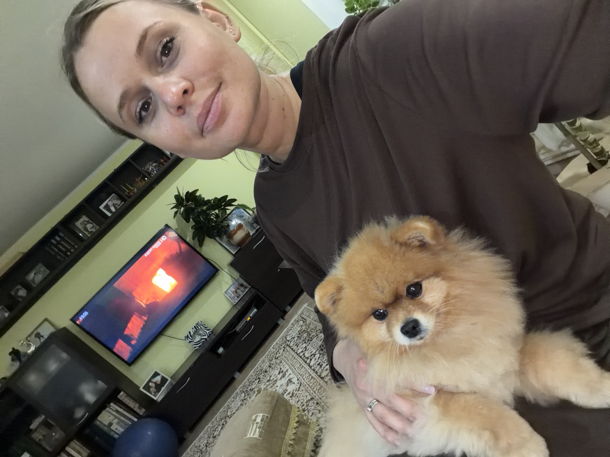 A woman holds a Pomeranian dog. A television is on a shelf showing a film called "Дом кино HD" and it is 19:55, presumably in Sarapul, Russia.