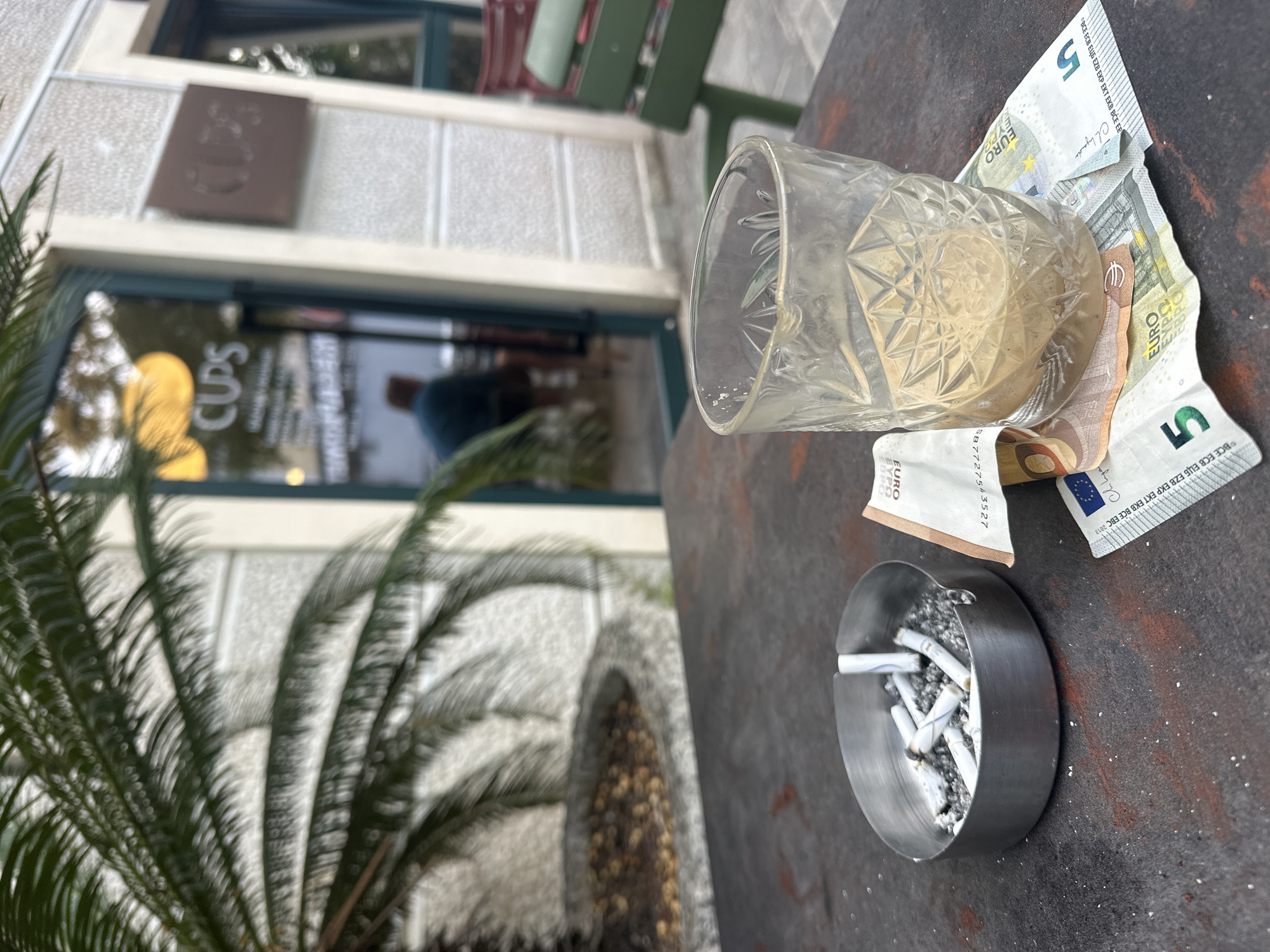 The image captures a scene at an outdoor café, likely in Budva, Montenegro, given the "CUPS" sign visible in the background and the Euro currency on the table. A partially-empty crystal glass sits on a worn metal table, surrounded by Euro banknotes, including 5 Euro notes. An ashtray overflowing with cigarette butts rests near the edge of the table. In the background, a blurred view of the café's entrance, a potted plant, and a glimpse of the building's architecture can be seen. The overall setting is relaxed but untidy, suggesting recent activity.