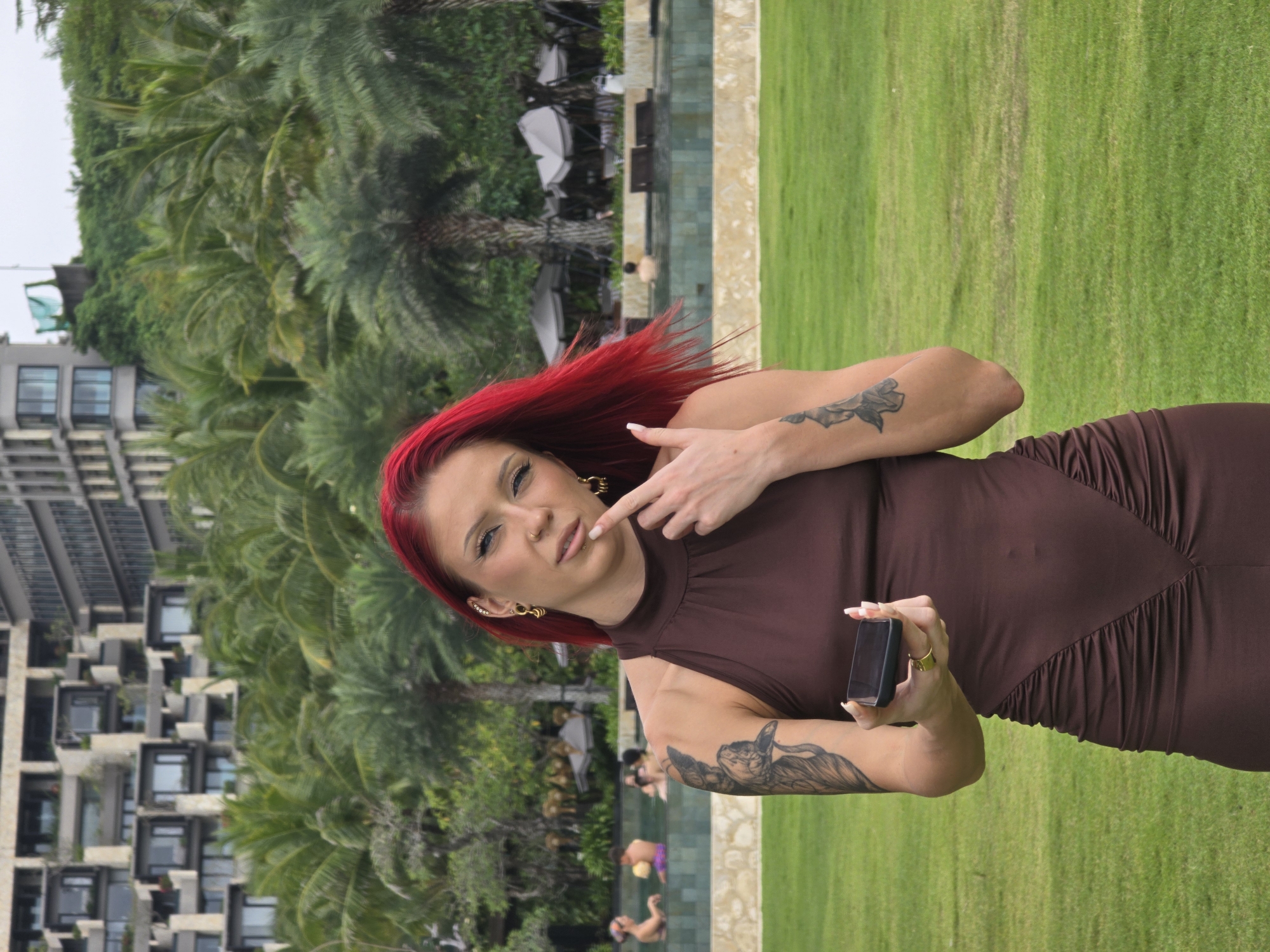 A woman with bright red hair stands on a grassy lawn. She is wearing a tight-fitting, dark brown dress and has tattoos on both arms. She holds a smartphone in her right hand and points to her lips with her left index finger. In the background, there is a multi-story building with palm trees and lush greenery. A swimming pool with people in it is also visible. The location is Nusa Dua, Indonesia, indicated by the tropical environment and architecture.
