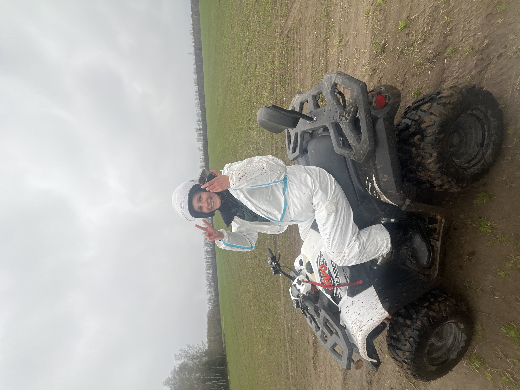 The image depicts a person in a white protective suit and helmet sitting on a white and black ATV. They are giving a peace sign. The ATV is dirty with mud splatters. The background shows a field with trees in the distance, under a cloudy sky. The location is Раздолье, Russia.