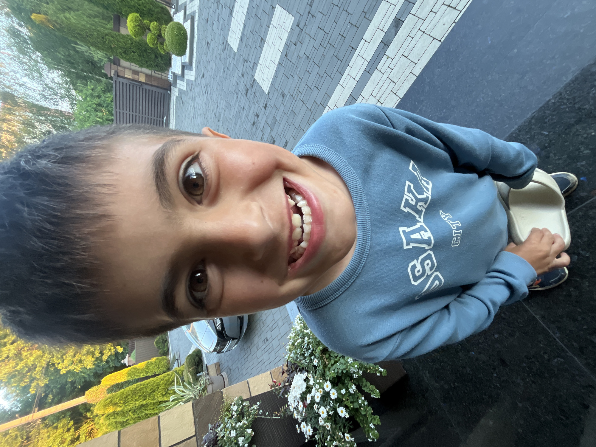 A young boy with dark hair and brown eyes is standing outside, looking directly at the camera with a wide, toothy smile. He is wearing a blue sweatshirt with white lettering that reads "OSAKA CITY." He is holding a light-colored object in his hands. Behind him, a paved walkway with a geometric pattern is visible, along with a car, various plants, and trees. The scene appears to be a residential area, possibly in Koroviia, Ukraine.