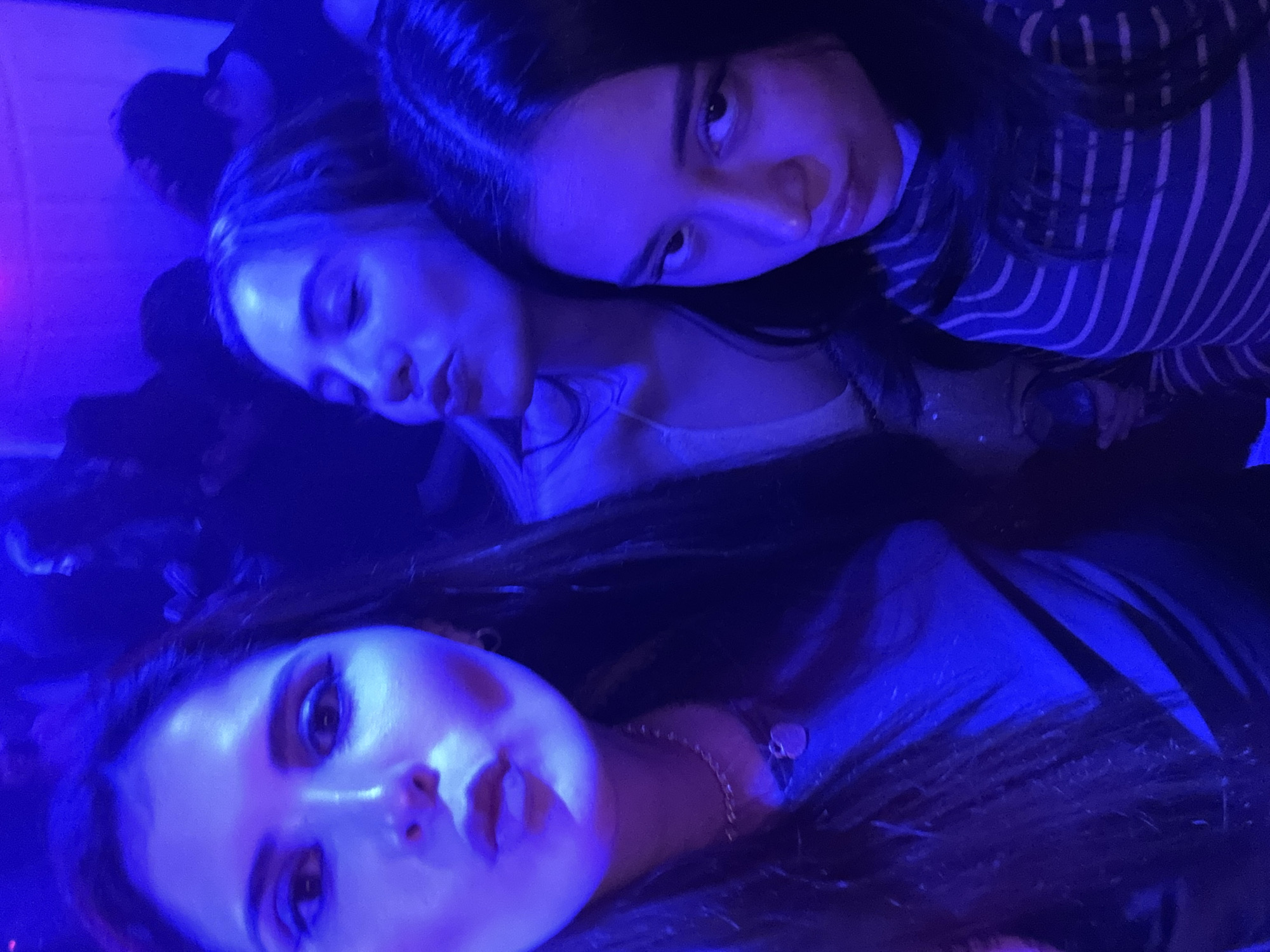Three young women pose close together, illuminated by blue lighting. The woman at the bottom of the frame looks directly at the viewer. The other two women are positioned above, one with pursed lips and the other looking directly at the viewer. They are indoors. There is no location information available in the image.