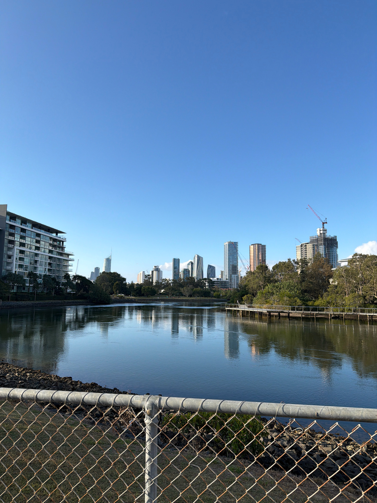 The image shows a wide, calm body of water reflecting a clear blue sky and a distant city skyline. On the left, a multi-story apartment building with balconies overlooks the water. Trees and vegetation line the opposite bank, interspersed with various modern buildings and construction cranes, indicating ongoing development. A wooden boardwalk extends over the water on the right side of the frame. In the foreground, a chain-link fence with a metal pipe top rail runs horizontally across the lower portion of the image, obscuring the immediate bank of the water. The scene appears to be a waterfront area within a city, likely Broadbeach Waters, Australia, given the architectural style and lush greenery.