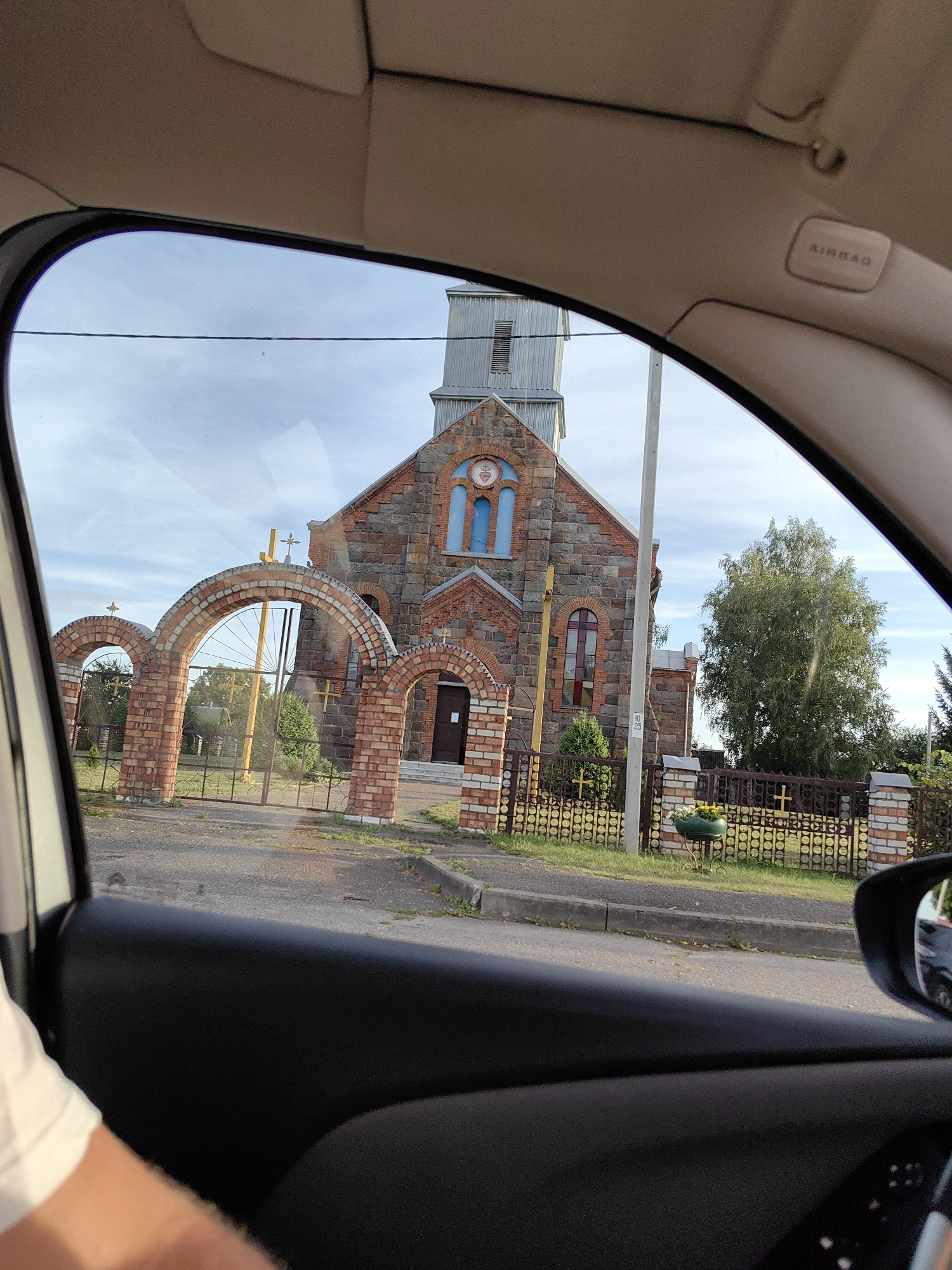 The image shows a view from inside a car, looking out at a stone church with arched brick gates and a fence. The church has a tall, metal-clad tower. The sky is partly cloudy and blue. The gates are made of brick with arched openings, and a metal gate is visible behind the central arch. A metal fence with circular patterns runs along the property line. A large tree is visible to the right of the church. The interior of the car is partially visible, including the dashboard and a car door. The location is identified as Ilyanskiy selskiy Sovet, Belarus.