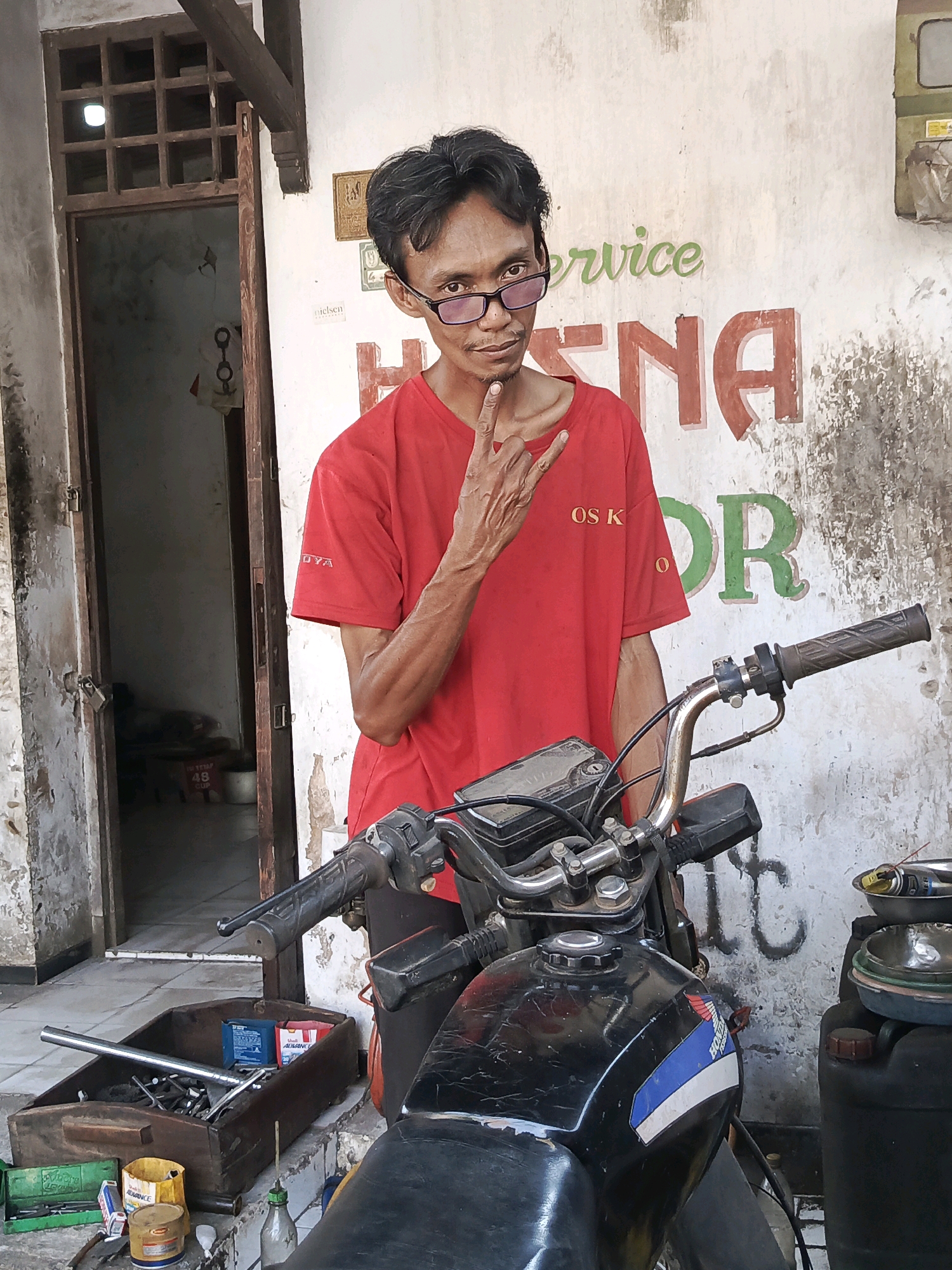 A man in a red t-shirt and glasses stands with a motorcycle in front of a weathered building with a painted sign reading "SERVICE HASNA MOTOR." The building's wall is rough and stained. To the left, a doorway leads into a dark interior. To the right of the motorcycle, a large black fuel can is visible. In the foreground, a wooden toolbox filled with tools, including a silver metal rod, is on the ground. The man is looking towards the camera and making a hand gesture. The scene suggests a mechanic's workshop in Semanding, Indonesia.