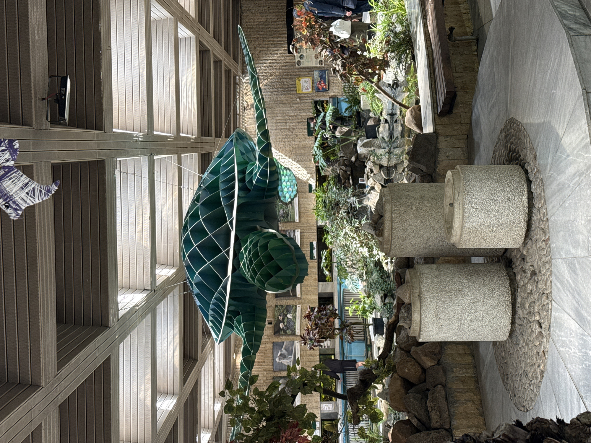 The image captures an indoor scene featuring a large, abstract sculpture of a green sea turtle suspended from the ceiling. The sculpture has a geometric, wire-frame design.
In the background, there is a faux waterfall constructed with rocks and various plants. Several cylindrical stone benches are also present near the waterfall, positioned on a stone-covered base.
The background also includes a wall with framed images. A section of the image shows a person standing near the plants in the background.
The location is Sofia, Bulgaria.