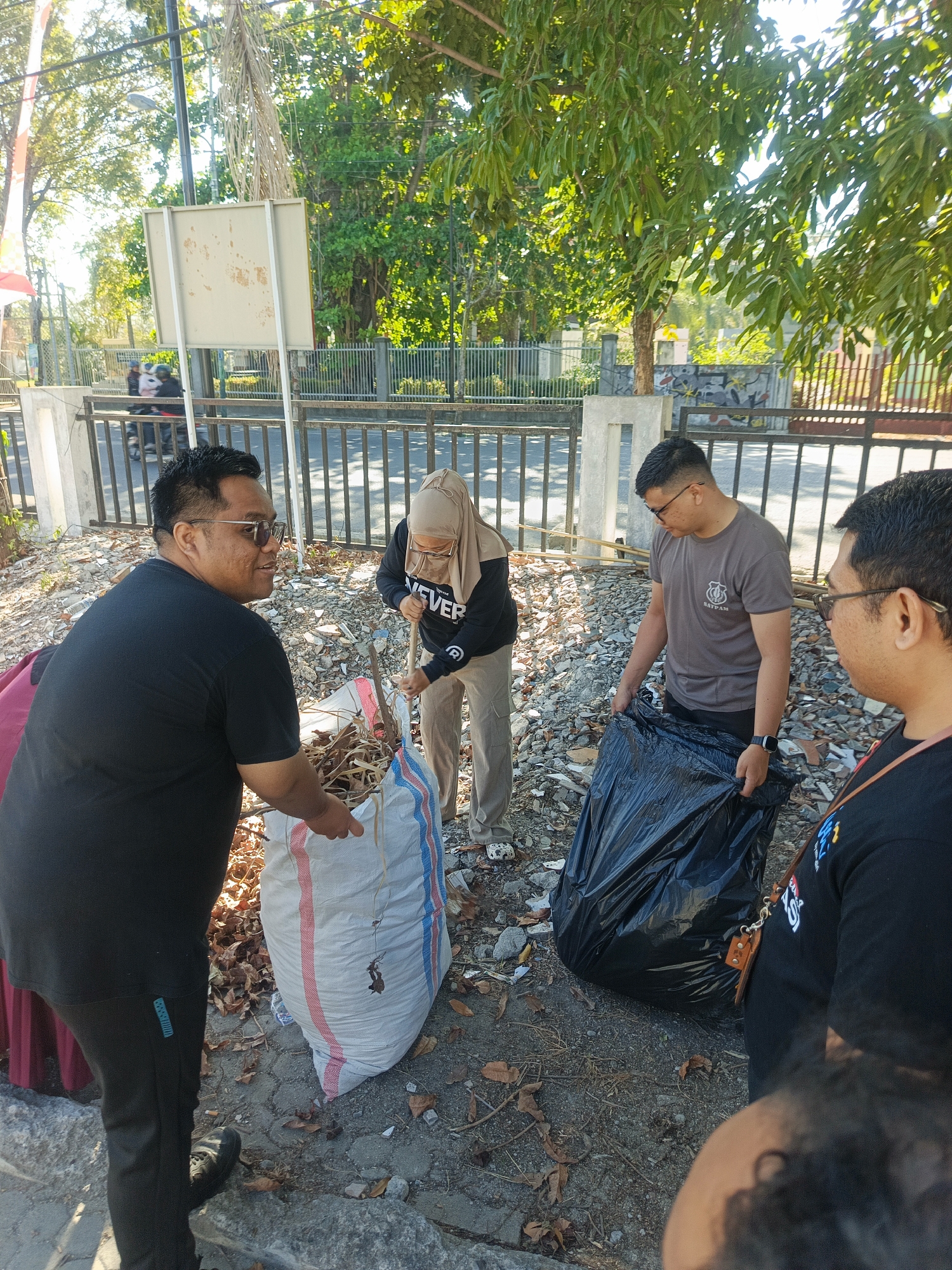 The image shows four individuals engaged in cleaning up debris on a sunny day in Gorontalo, Indonesia. In the foreground, a man wearing a black t-shirt and sunglasses is holding a large, white sack filled with dry leaves and other organic matter. To his right, a woman in a hijab and a black hoodie is using a wooden stick to gather more debris into the sack. Further to the right, a man in a grey t-shirt, identified by the "SATPAM" logo, is holding a large black plastic bag, ready to collect more waste. Another person, only partially visible in the foreground on the right, is also observing the activity.
The environment appears to be an outdoor area, possibly a roadside or public space, with a metal fence and some trees in the background. The ground is covered with scattered leaves, rocks, and what looks like construction debris. Sunlight is casting shadows, indicating it's likely late morning or early afternoon. The presence of the "SATPAM" logo on one of the shirts suggests a security or public order context. The overall scene depicts a community effort to clean up a public area in Gorontalo.