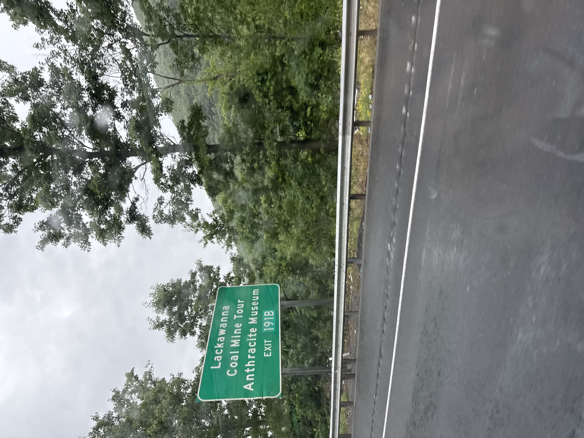 A green highway sign with white lettering reads "Lackawanna Coal Mine Tour Anthracite Museum" and "EXIT 191B". The sign is positioned in front of a dense forest of green trees under a cloudy, overcast sky. A wet asphalt road with a white lane marker stretches across the foreground, indicating recent rain. A metal guardrail runs parallel to the road, separating it from the wooded area behind. The overall scene is that of a roadside view in a rural or semi-rural area, likely in South Abington Township, Pennsylvania, given the mention of Lackawanna County on the sign.