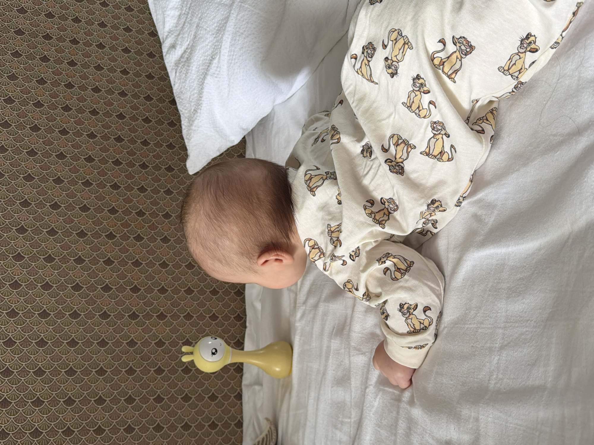 A baby wearing pajamas patterned with Simba from "The Lion King" is lying on a white bed next to a white pillow. Beside the baby is a yellow giraffe-shaped toy. The bed is placed in a room with a patterned brown and beige floor. This photo was taken in Khimki, Russia.
