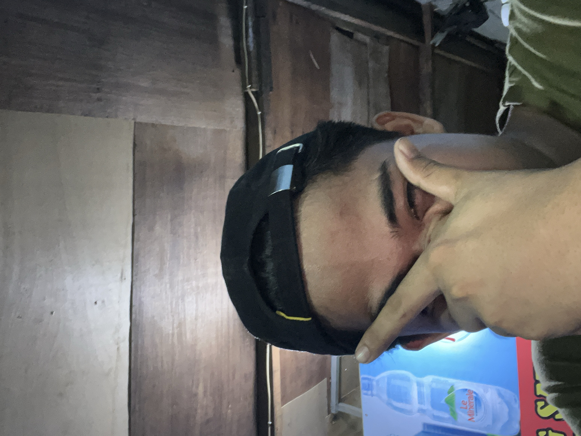 A low-angle medium shot captures a person wearing a black baseball cap, partially obscuring their face with their right hand. The person's eyes are visible beneath the brim of the cap. The background consists of a wooden ceiling. In the lower left corner, a portion of a blue advertisement featuring a water bottle and the text "Le Minerale" is visible. The scene appears to be indoors, possibly in Central Jakarta, Indonesia, given the presence of the "Le Minerale" branding, a popular bottled water brand in Indonesia. The lighting is dim, with a visible light source reflecting on the wooden ceiling. The overall impression is casual and informal.