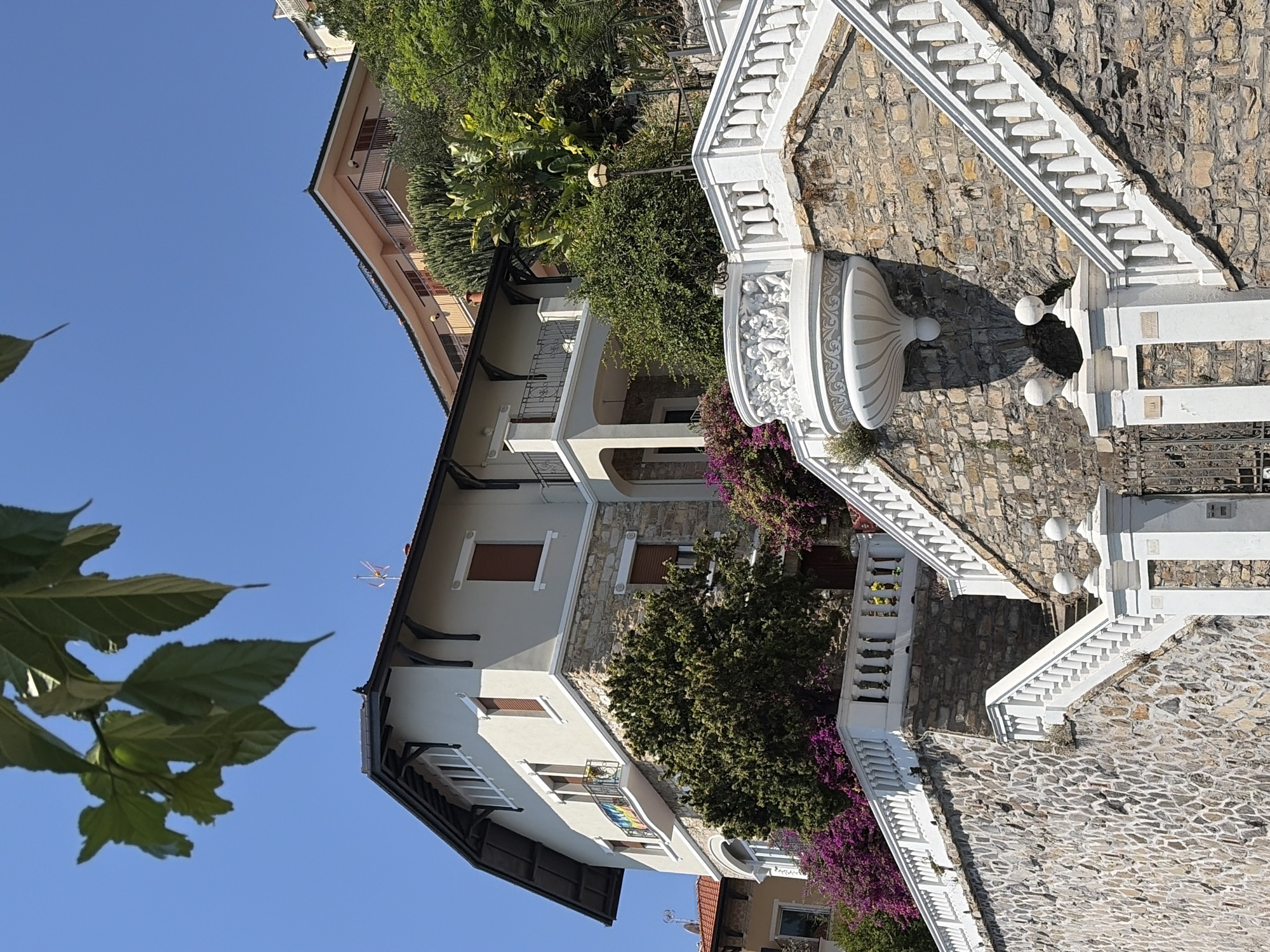 This is a medium shot, outdoor, eye-level, vertical photograph taken on a clear day. The image features a multi-story house built on a hillside, with a prominent stone-clad staircase leading up to it. The house itself is a cream color with dark trim and a dark roof, featuring stone accents around its base. There are balconies with railings on the upper floors, and dark brown shutters on the windows. Lush green trees and vibrant purple flowers are visible surrounding the house and along the staircase.
To the right of the main house, a section of another building with a peach-colored facade and balconies can be seen. The stone staircase is wide and elaborate, with white balustrades and decorative elements, including a sculpted white fountain feature with cherubic figures. The lower part of the staircase is flanked by a substantial stone retaining wall. A black metal gate is visible at the bottom of the staircase.
Based on visual cues such as the architecture, the Mediterranean vegetation, and the presence of a rainbow flag on the balcony of the main house (suggesting a possible pride event or location), this scene is likely located in the Liguria region of Italy, specifically the city of Imperia. The overall impression is one of a luxurious, well-maintained property in a sunny, coastal environment. There are no people or animals visible in the image.