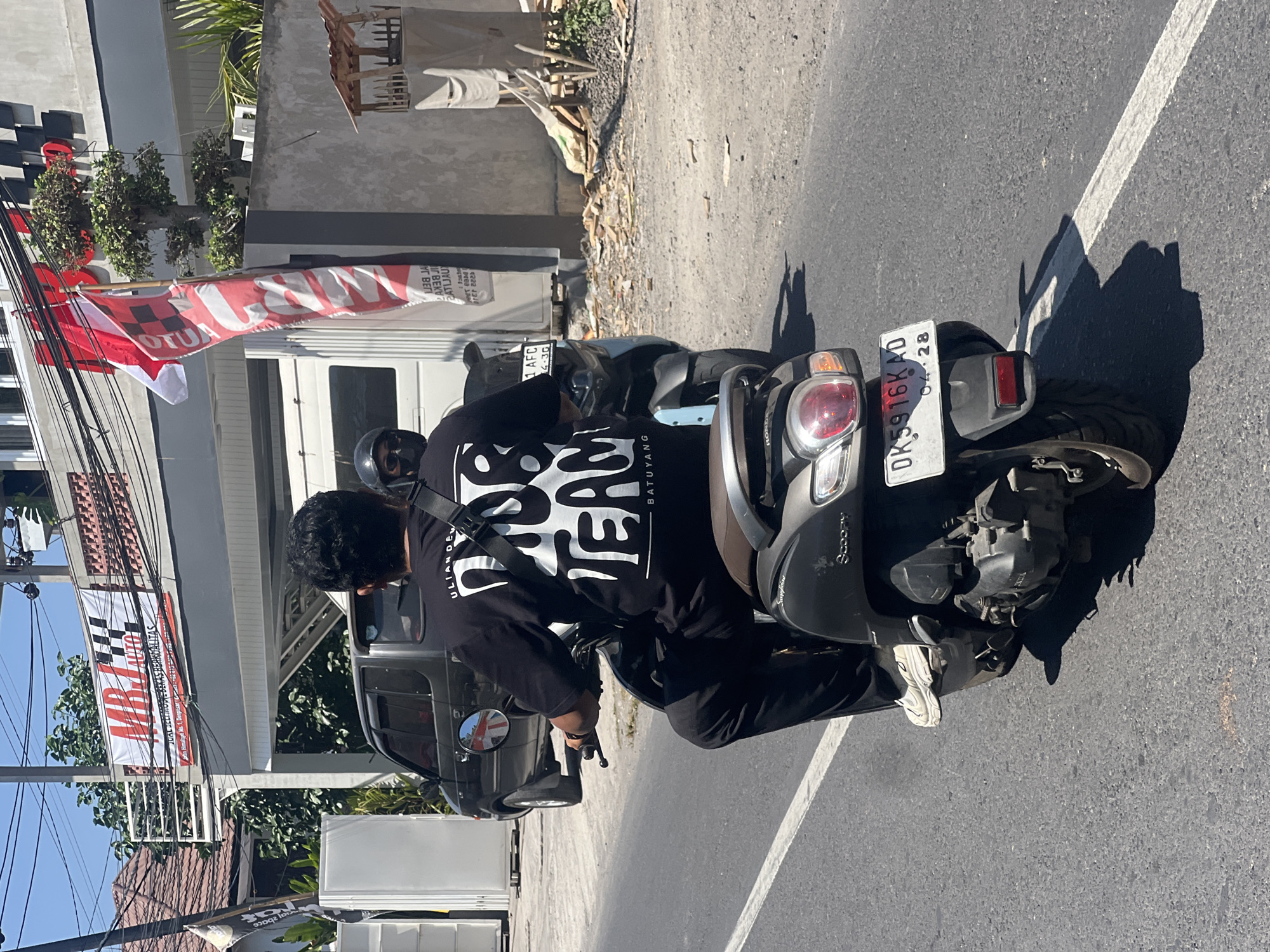 In Denpasar, Indonesia, a person rides a dark gray scooter down a paved road. The rider, wearing a black t-shirt with white lettering that reads "ULI ANDEV" and "OUR TEAM," and black pants, is seen from behind. They are also wearing a helmet, sunglasses, and white sneakers. The scooter has a white license plate with the characters "DK5916KD" and "04-28" displayed. In the background, a building with a red and white banner that says "MBJ AUTO" is visible. Red and white flags are also flying near the building. Several cars are parked on the side of the road. The sky is clear and blue, suggesting it is a sunny day.