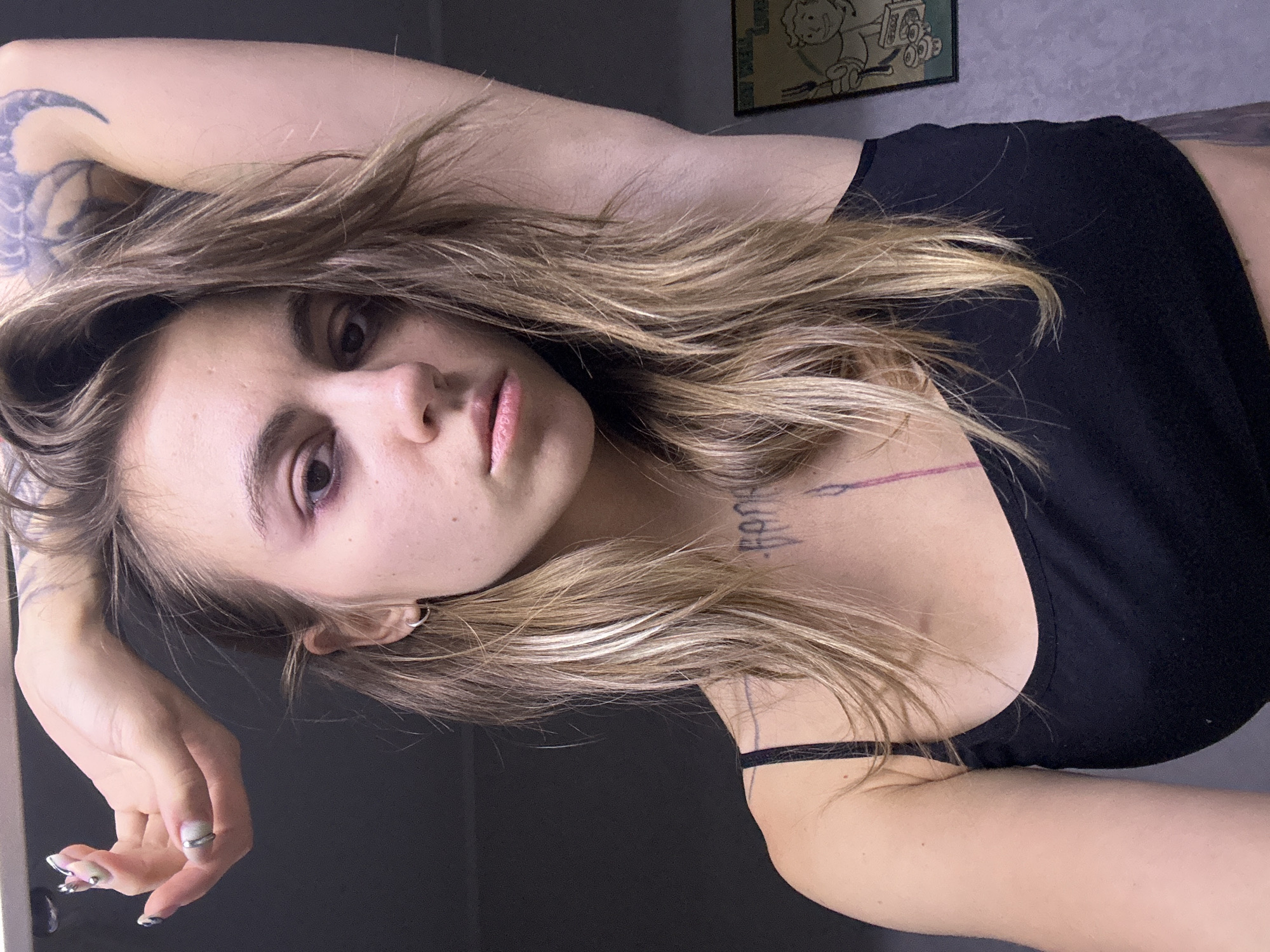 A young woman with long, blonde, messy hair is posing for a selfie in what appears to be a dimly lit room with a neutral gray wall in the background. She is wearing a black tank top. Her right arm is raised above her head, and her left hand is resting on her head. She has visible tattoos on her arm and neck. Her makeup includes dark eye shadow and eyeliner. A framed picture of the character "Vault Boy" from the Fallout video game franchise is visible on the wall to the right. There is no discernible information in the image to suggest a location in Moscow, Russia.