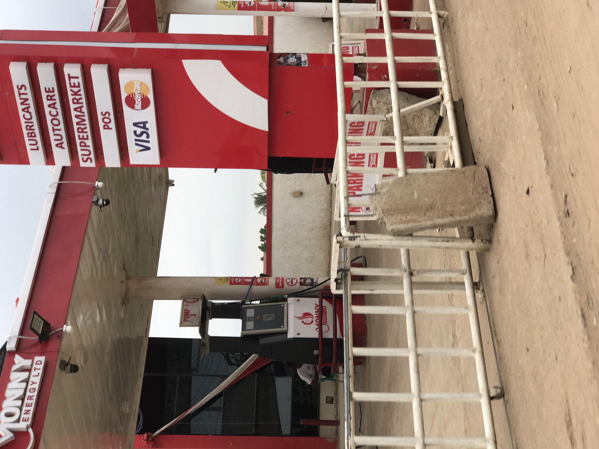 The image depicts a gas station in what is likely Abuja, Nigeria, identifiable by the "MONNY ENERGY LTD" branding. The station offers lubricants, autocare, and supermarket services, accepts Visa and Mastercard, and has at least one pump labeled "Pump 3." A white metal barrier sits in front of the gas pumps. The environment appears urban or peri-urban.