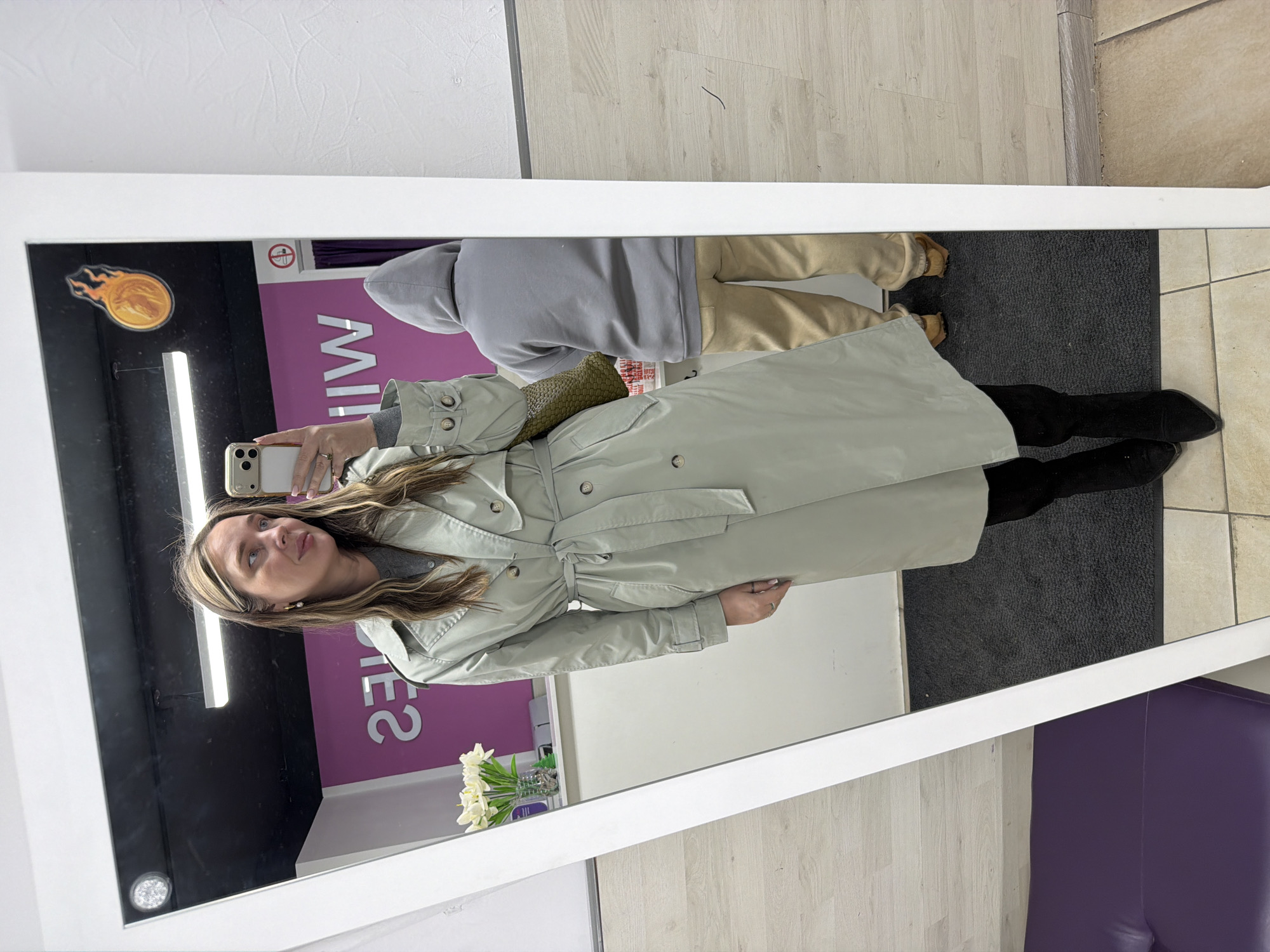The image displays a full-length mirror selfie taken by a woman indoors, likely within a retail or commercial establishment in Moscow, Russia. The woman is positioned centrally, holding a light-colored smartphone with multiple cameras to capture her reflection. She has long, highlighted brown hair and is wearing subtle earrings and a ring.
Her attire consists of a stylish, light sage green or pale grey trench coat with buttons and a belted waist, worn over a grey top. She is also wearing dark, possibly suede, knee-high boots. A green, textured or quilted bag hangs over her left shoulder. She looks slightly upwards and to the side, maintaining a neutral expression.
The setting features a large mirror with a white frame. The floor consists of light wooden laminate transitioning to a light-colored tiled section, with a black mat placed in between. Fluorescent strip lighting illuminates the space.
In the reflected background, behind the woman, a second individual is partially visible. This person is wearing a grey hooded sweatshirt, light brown or khaki pants, and light-colored slip-on shoes, possibly moccasins. This reflection is mostly obscured by the main subject and the mirror frame.
A distinctive purple wall or partition is reflected in the background, adorned with white, mirrored text that appears to read "MIW" and "IES," likely part of a brand name or signage. A "No smoking" sign icon is also visible in the reflection. To the right of the woman, a small vase with white flowers rests on a counter. A flaming comet or meteor sticker in orange and yellow is adhered to the upper left corner of the mirror. A purple upholstered bench or seat is partially visible in the lower right foreground reflection. The overall scene suggests a casual moment of self-documentation in a well-lit, public indoor environment.