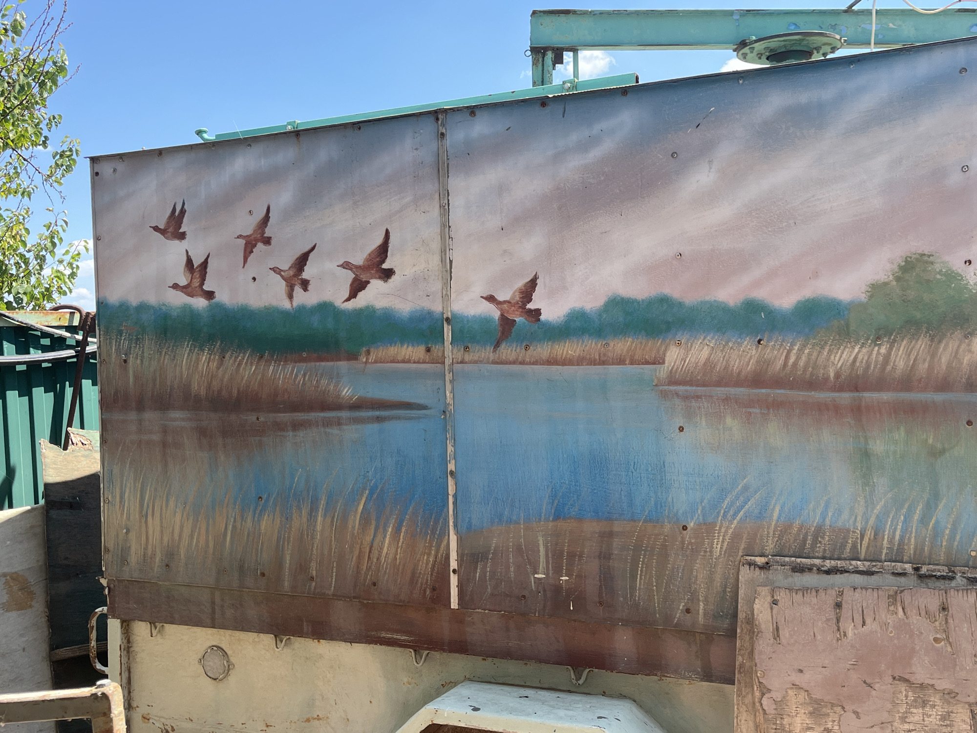 The image shows a mural painted on the side of a weathered trailer, depicting a flock of ducks in flight over a body of water with reeds and trees in the background. The sky above is a clear blue. The mural is on a metallic surface, likely the side of a utility vehicle or storage container, with visible rivets and seams. The trailer itself appears to be in a state of disrepair, with rust and peeling paint evident. In the background, part of a green metal fence and some foliage are visible. The scene suggests an outdoor setting, possibly in a rural or semi-industrial area.