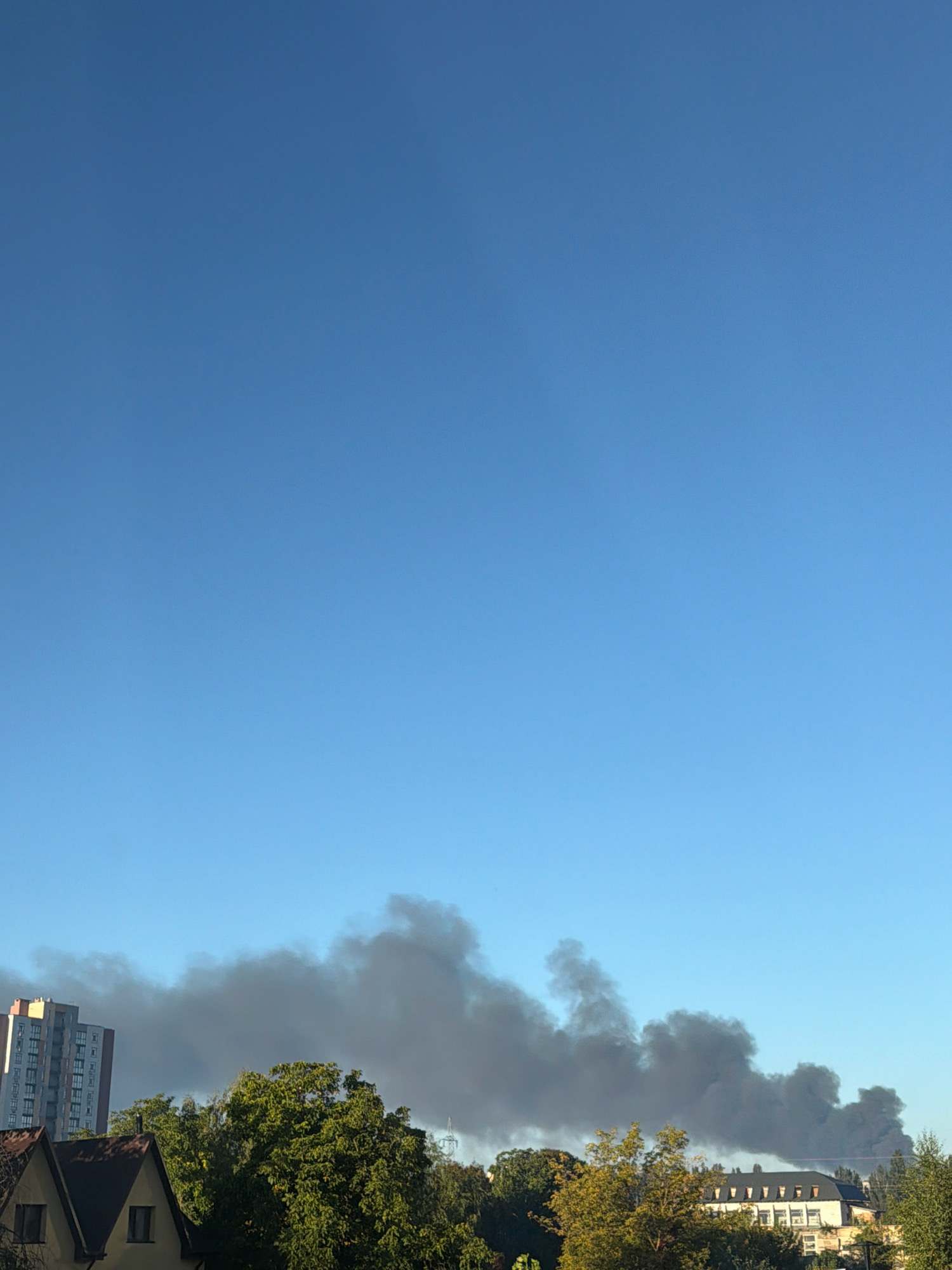 The image shows a clear blue sky over a city with some buildings and trees. Thick black smoke billows upwards and across the sky, obscuring parts of the buildings and trees. On the left, a tall, modern building with many windows is visible. In the foreground, there are rooftops of houses and lush green trees. Further to the right, a long, white building with multiple windows and a dark roof is partially visible behind the trees. The presence of smoke suggests an incident, possibly a fire, occurring within the city. The scene appears to be captured during daylight, given the bright sky and the illumination of the buildings.