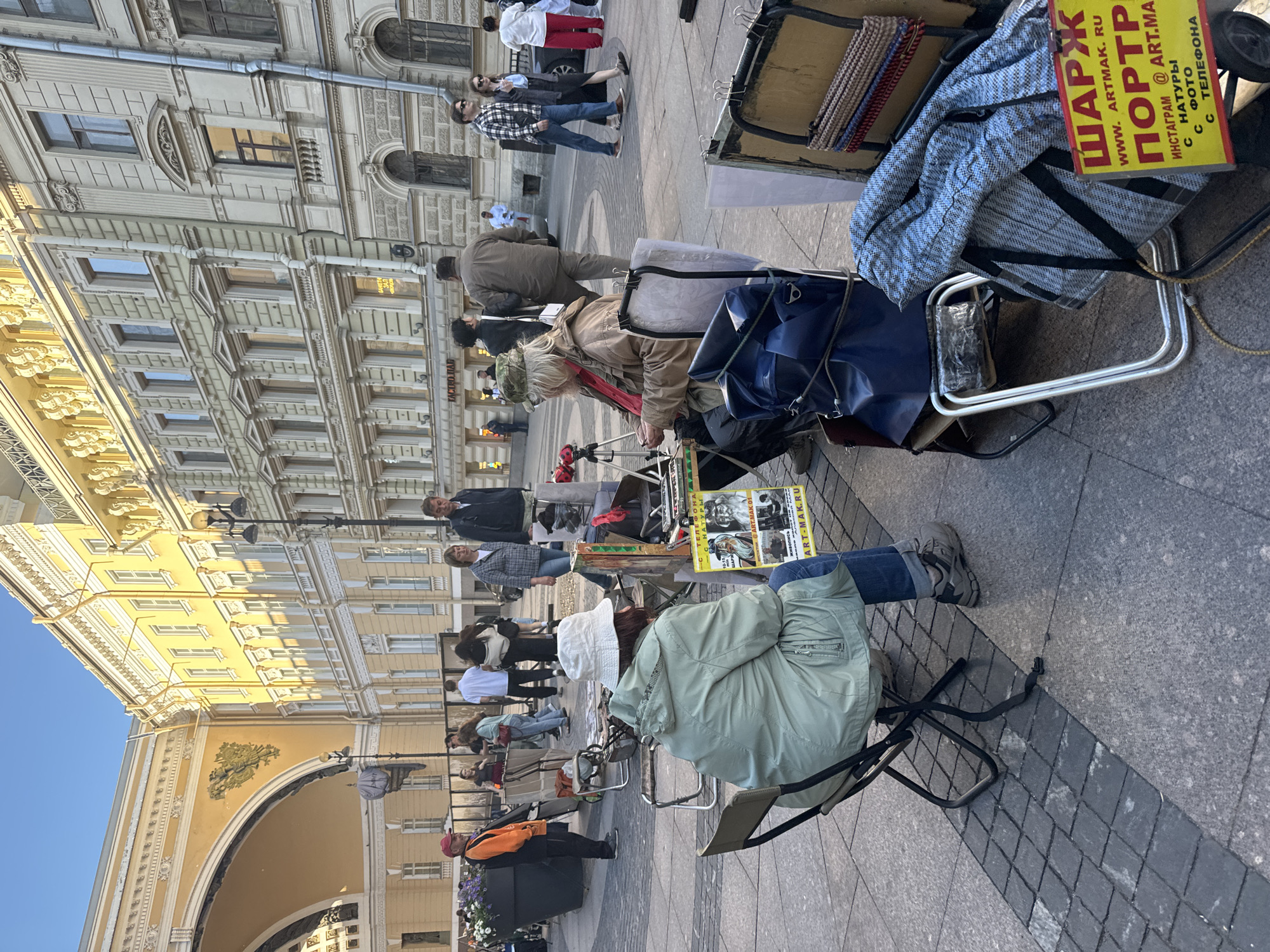 This image captures a street scene in Saint Petersburg, Russia, likely in Palace Square, given the architectural style of the buildings, particularly the yellow building with ornate detailing and the large archway. The foreground features two individuals, presumably artists, set up outdoors.
On the left, a person wearing a light green hooded jacket and a white bucket hat sits in a folding chair, facing away from the camera. They are seated in front of a table displaying what appears to be artwork or photographs. In front of this individual is a sign written in Russian, featuring yellow and red lettering, with the text "ШАРЖ" (which translates to "Caricature") and "ПОРТР" (likely short for "Портрет" or "Portrait"). Below this, there is additional text including "WWW.ARTMAK.RU" and an Instagram handle.
To the right of this person, another individual, wearing a tan jacket and a camouflage hat, is also seated and appears to be engaged in drawing or painting, with a tripod and art supplies visible.
The background shows a busy urban environment with more people walking by. The buildings in the background are of classical architecture, with numerous windows and decorative elements. The overall atmosphere suggests a pleasant day, with clear blue skies and sunshine casting shadows on the paved ground. The scene is typical of outdoor art markets or street artists found in prominent public spaces.