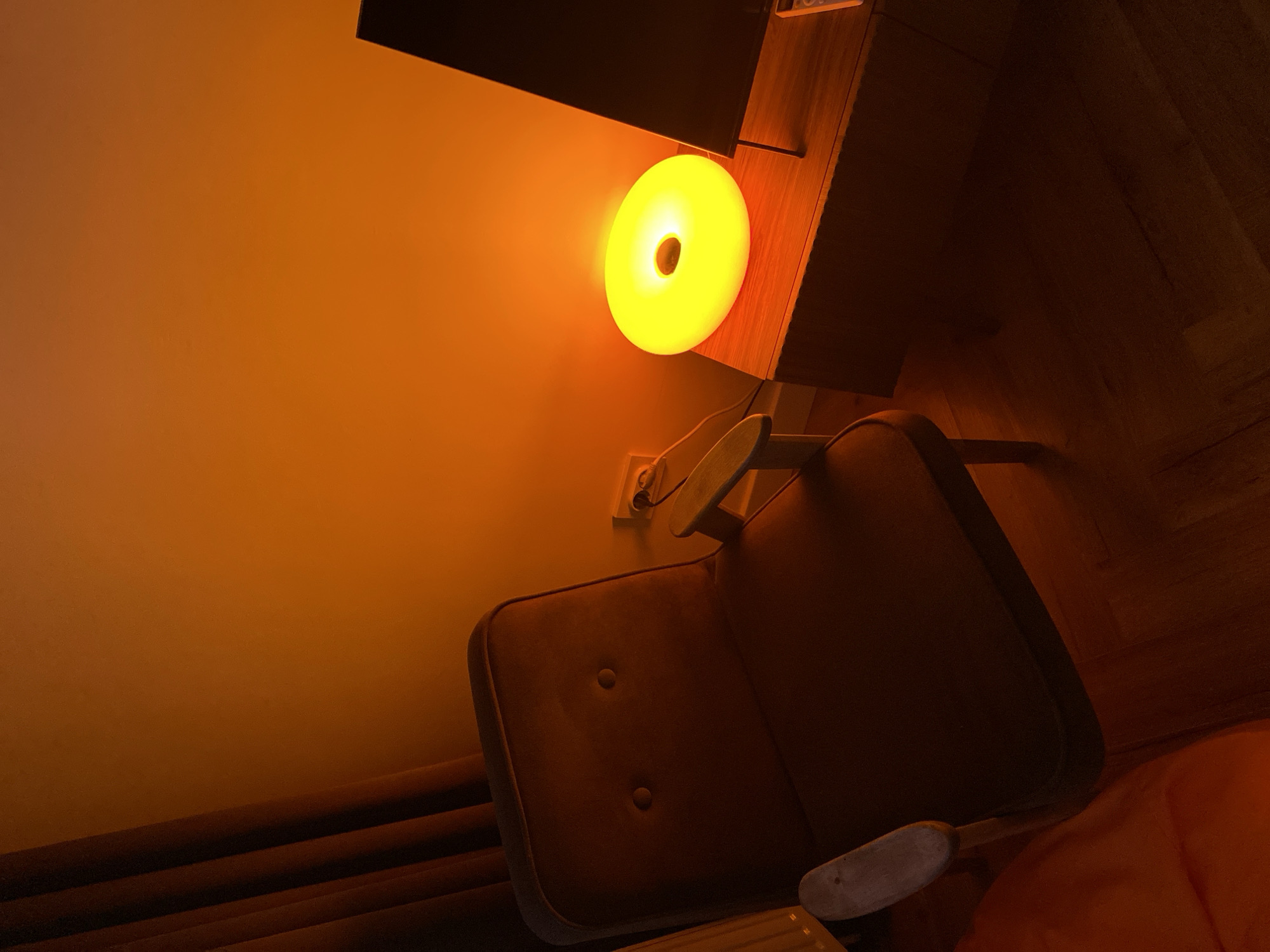 The image depicts a dimly lit interior room, likely in the evening. A brown, upholstered armchair with two buttons is situated near a desk or shelf. A circular, yellow light source is attached to the desk, illuminating the wall behind it. An electrical outlet is visible on the wall near the chair. The scene appears to be in a domestic setting, though the specific location in Izhevsk, Russia, cannot be determined from the visual information alone.