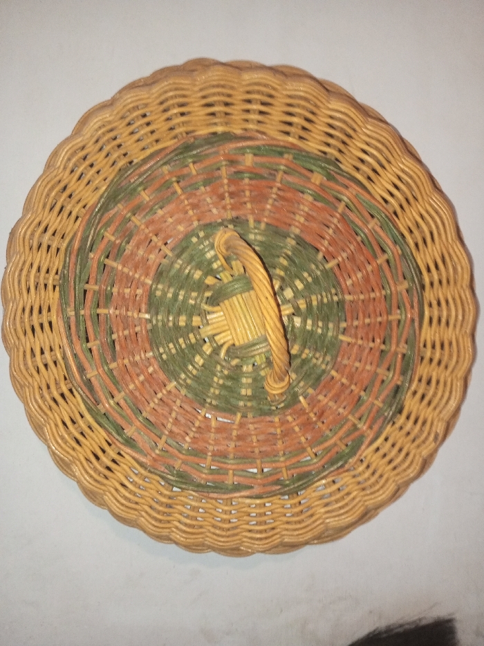 A round wicker basket lid with a handle in the center. The lid has concentric circles of woven wicker in alternating colors of green and brown. The outer rim of the lid is a lighter tan color and features a scalloped edge. The handle is also woven in the same light tan wicker, secured in the center of the lid. The overall impression is of a handcrafted and rustic item.