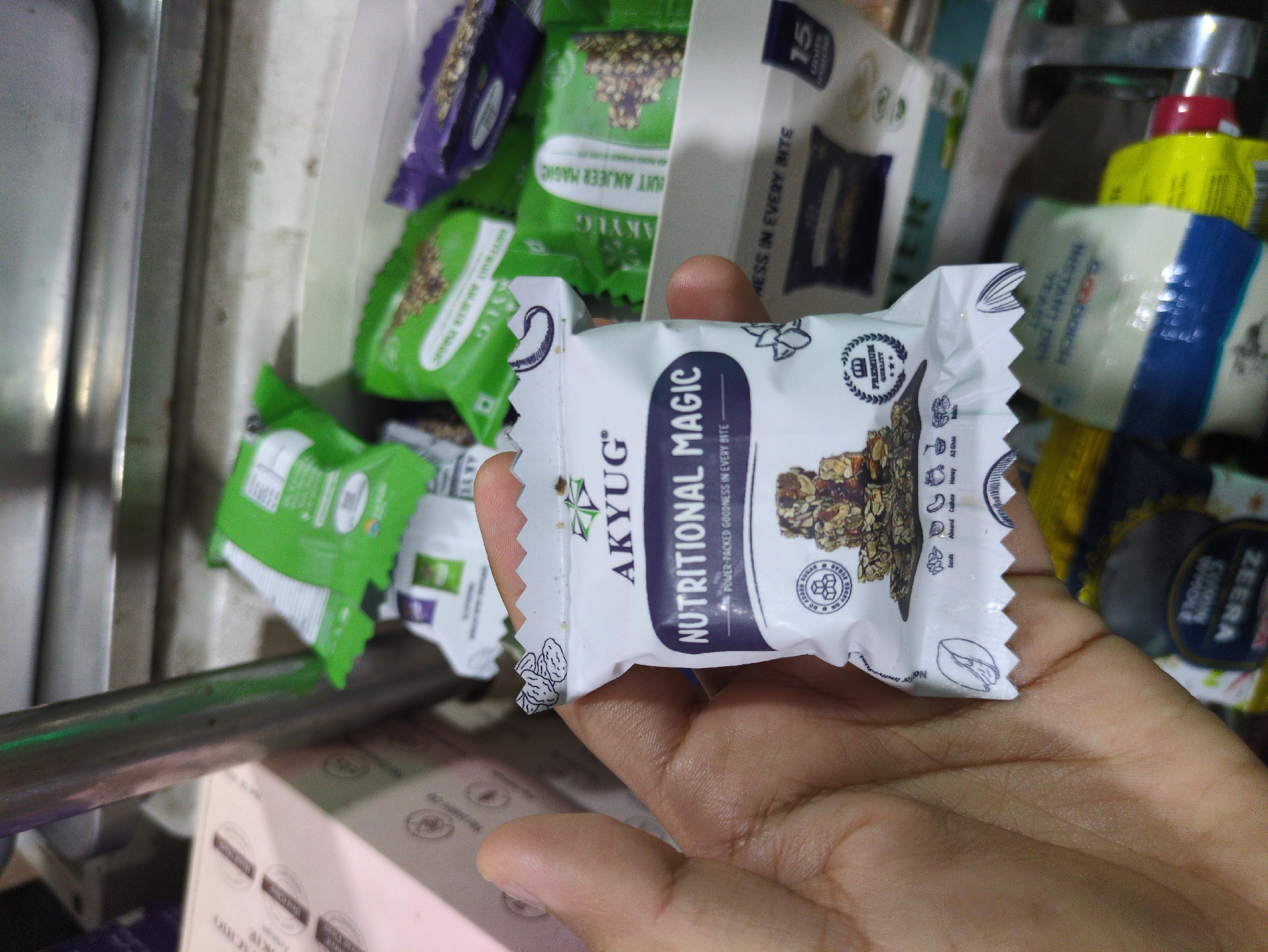 A hand is holding a small, individually wrapped snack bar, labeled "AKYUG NUTRITIONAL MAGIC POWER-PACKED GOODNESS IN EVERY BITE." The packaging is white with dark blue and green accents. The snack bar itself, visible through a clear window, appears to be a mix of nuts and seeds bound together. In the background, other similar snack bars are visible in green and purple packaging, some stacked in a container. The scene appears to be in a shop or market stall, possibly in Bhagsu, India, given the context provided by the surrounding objects and packaging, though this cannot be definitively confirmed from the image alone. The lighting is somewhat dim, suggesting an indoor environment.
