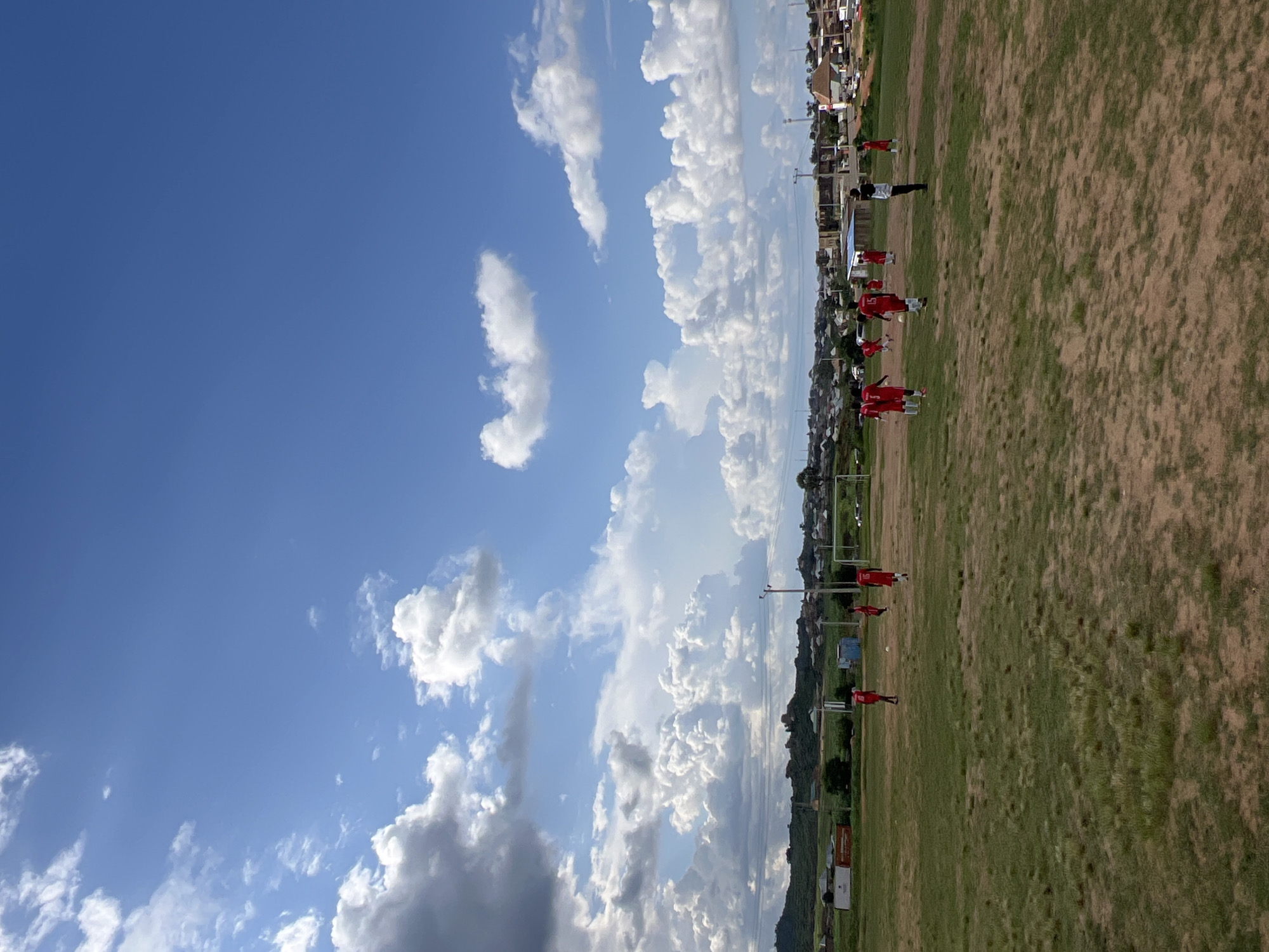 The image depicts an outdoor football game taking place on a dusty, uneven grass field under a partly cloudy blue sky. A group of individuals, mostly wearing red jerseys, are engaged in playing football. One person in a black and white shirt stands observing the game. In the background, a town or city with numerous buildings is visible, situated on a hilly landscape. The scene appears to be in Jos, Nigeria, given the context provided. The overall environment suggests a rural or semi-urban setting with the presence of a sports field and a populated area in the distance.