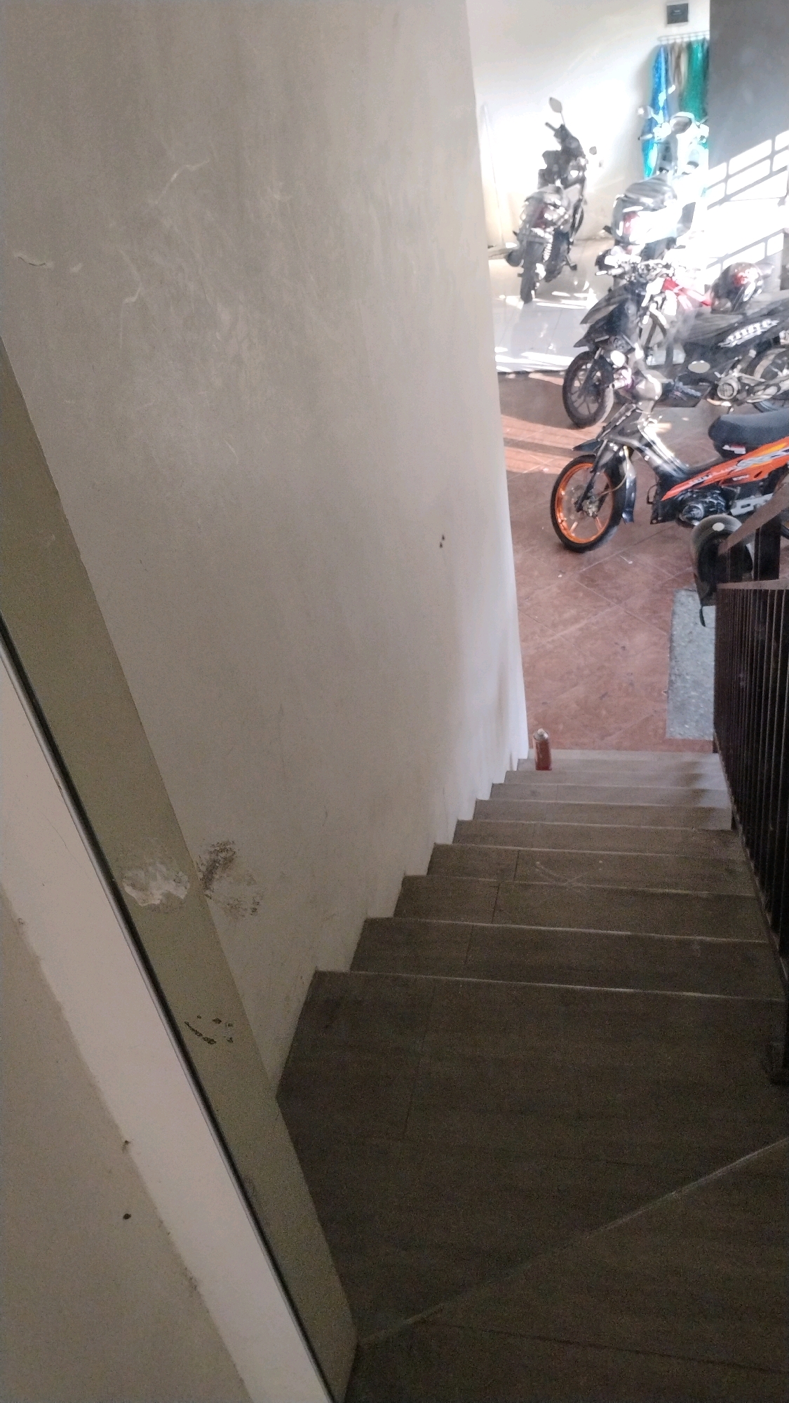 The image shows a dimly lit, indoor stairwell with a wall on the left and a railing on the right. Several motorcycles are parked in the background, visible through an opening to another area. The floor of the stairwell is tiled, and a single red object, possibly a bottle, is visible on one of the steps. The wall on the left appears to be concrete and shows some signs of wear and tear. The overall impression is of a functional, perhaps slightly neglected, space. The location in Gumpang, Indonesia is not explicitly discernible from the visual elements alone, but the presence of multiple motorcycles suggests a residential or commercial area where such transportation is common.