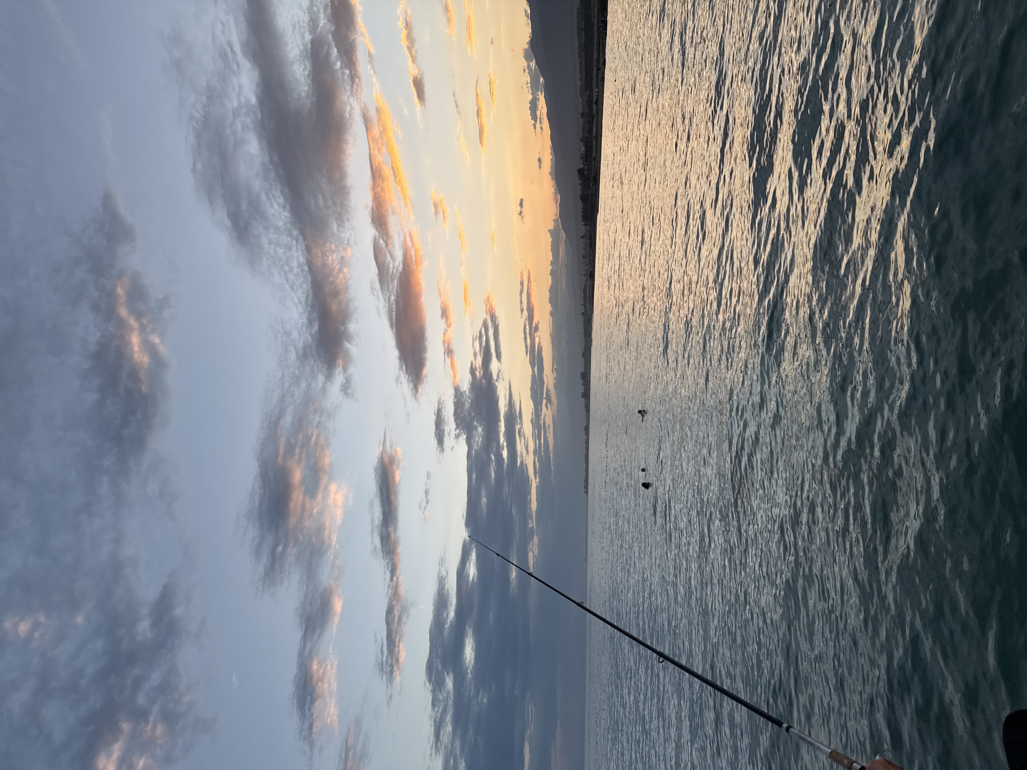 A fishing rod extends into the frame from the lower left corner, angled towards the right. The rod rests on the water's surface which is calm and rippled, reflecting the colors of the sunset. In the distance, several people are visible in the water, appearing to swim or float. The horizon is marked by a distant shoreline with buildings and mountains. The sky is a canvas of twilight colors, with shades of blue, gray, and pink from the setting sun creating a dramatic cloudy sky. The location is Sary-Oy, Kyrgyzstan.
