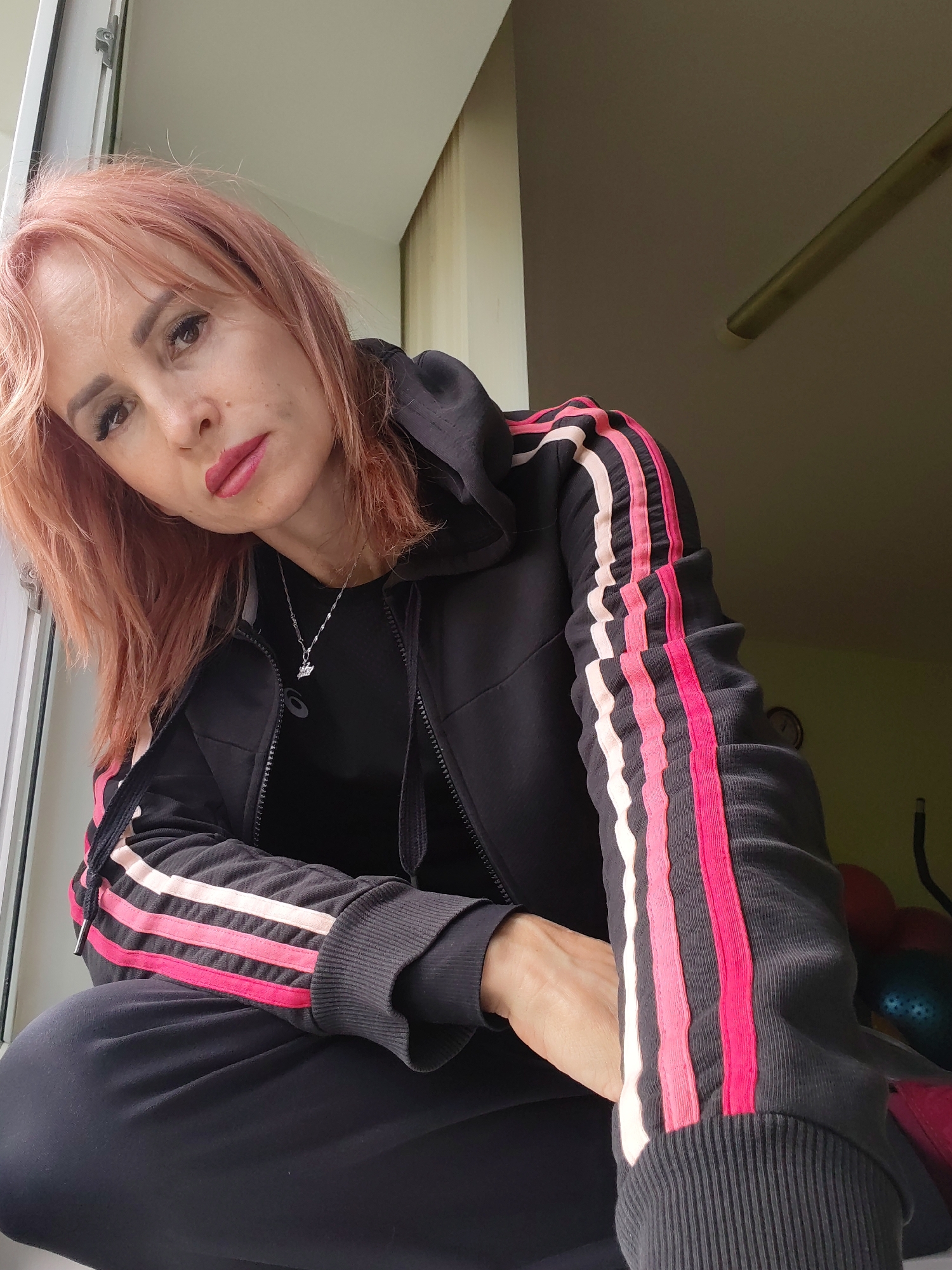 A woman with reddish-blonde hair and full makeup, wearing a black tracksuit with pink and white stripes on the sleeves, poses in what appears to be an indoor setting near a window. She is looking towards the camera with a slight tilt of her head. The background is dimly lit and shows a wall and a ceiling with a light fixture or beam. The overall impression is a casual portrait taken indoors. There is no indication of the city of Chaikovsky, Russia in the image.