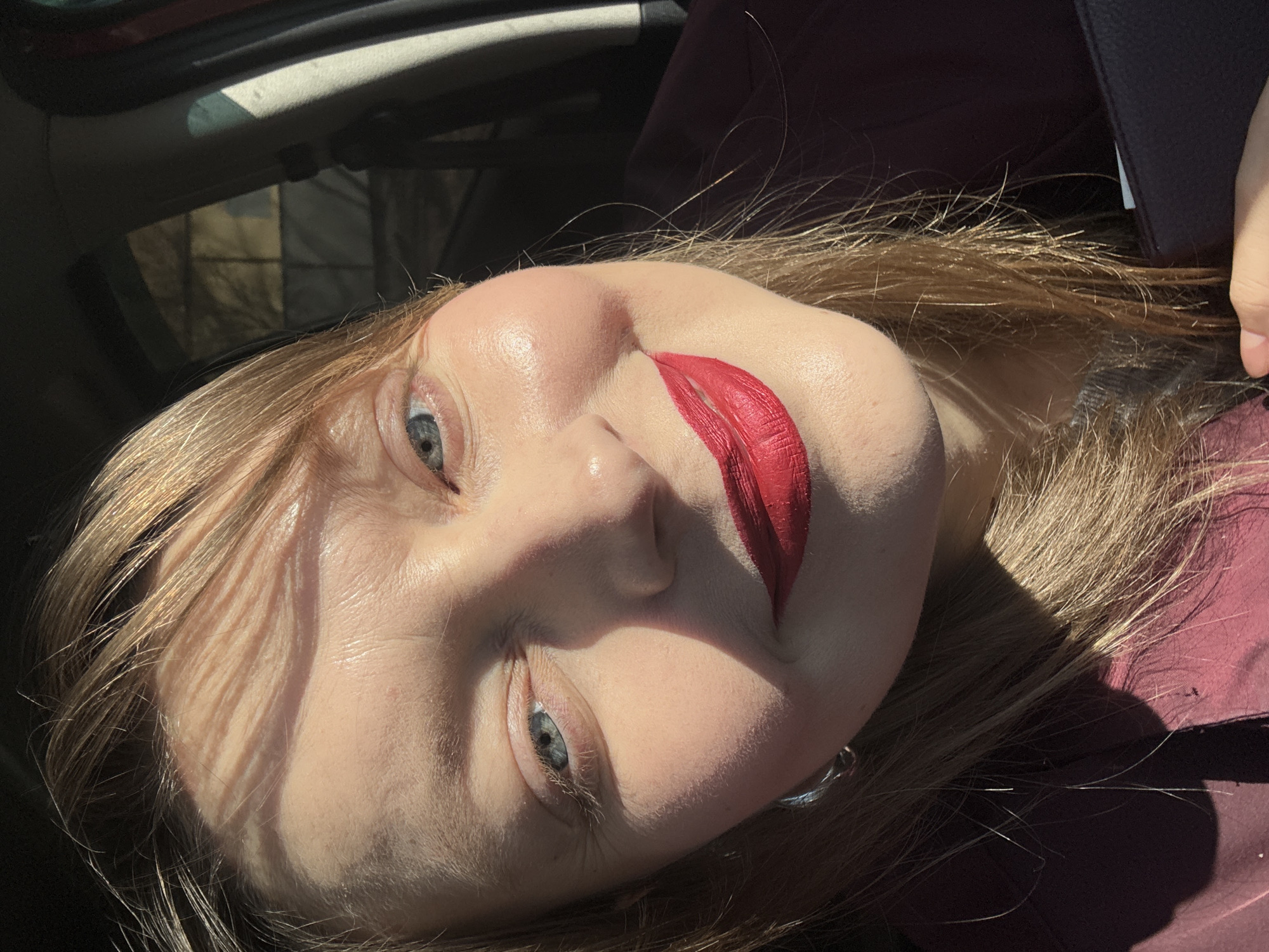 A close-up, rotated portrait captures a person, likely female, positioned within a car interior under strong sunlight. The subject has fair skin, light blue eyes, and long, light brown hair that reflects the bright illumination. A striking feature is the bright red lipstick, applied in a stylized, expressive manner that extends beyond the natural lip line, particularly on the upper lip, creating a dramatic aesthetic.
Harsh shadows and bright highlights are cast across the face, especially on the forehead, nose, and cheeks, indicating direct sunlight, suggesting daytime. The person gazes directly at the camera with a neutral to slightly contemplative expression. The car's interior appears dark, with glimpses of a roofline and reflections, while blurred shapes through a window hint at trees or structures outside. The subject is wearing a dark red or maroon garment. There is no visible text in the image. The location context for this media file is Izhevsk, Russia.