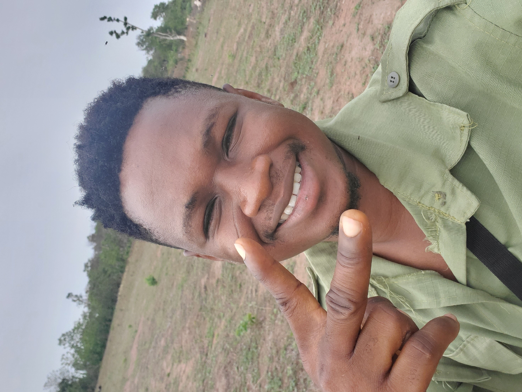 A young Black man is captured in a cheerful outdoor selfie or portrait, set in Mararaba, Nigeria. He is smiling broadly with his eyes squinting slightly, revealing white teeth. He has short, curly dark hair and light facial stubble. He wears a light olive green collared shirt, possibly a uniform or casual work attire, with visible stitching details and a button on a shoulder flap. His right hand is prominent in the foreground, raised to make a "peace sign" gesture with his index and middle fingers.
The background features a light grey, overcast sky, suggesting a cloudy day or soft daylight. Below the sky, there is a sprawling expanse of dry grass and dirt ground. In the mid-distance and further back, green trees and foliage line the horizon, indicating a rural or open natural environment. The man's expression conveys happiness and friendliness. There is no visible text in the image.