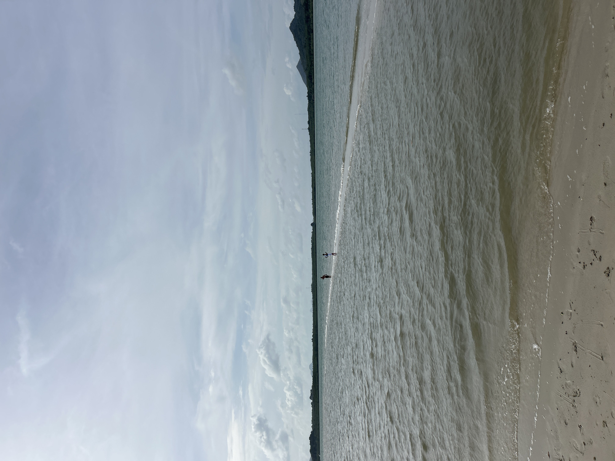 The image shows a wide, calm body of water in the foreground with a thin sandbar extending into it. Two individuals, appearing to be children, are walking on the sandbar. The water has small ripples and a slightly murky greenish-brown hue. The sandbar is a light beige color and is submerged in some areas by shallow water. In the background, there is a distant shoreline lined with dark green trees and vegetation, culminating in a dark, hilly landmass on the right. The sky above is overcast with diffused sunlight, featuring light gray and white clouds that are spread across the pale blue sky. The scene appears to be a natural coastal environment, possibly a beach or a shallow bay area. The location is identified as Ko Yao, Thailand, suggesting a tropical or subtropical setting.