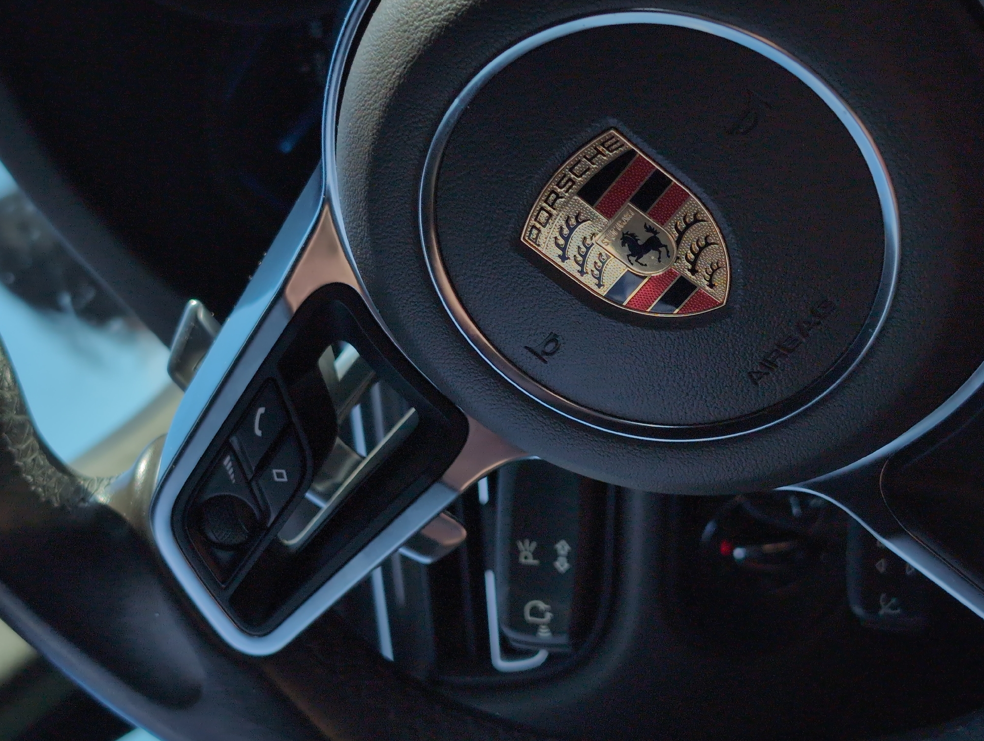 This image shows a close-up of a Porsche steering wheel, focusing on the center emblem and adjacent control buttons. The emblem is prominently displayed at the center of the steering wheel. To the left of the wheel are buttons. The location of this image is unknown, and there is no evidence of the city of Takarlikovo, Russia.