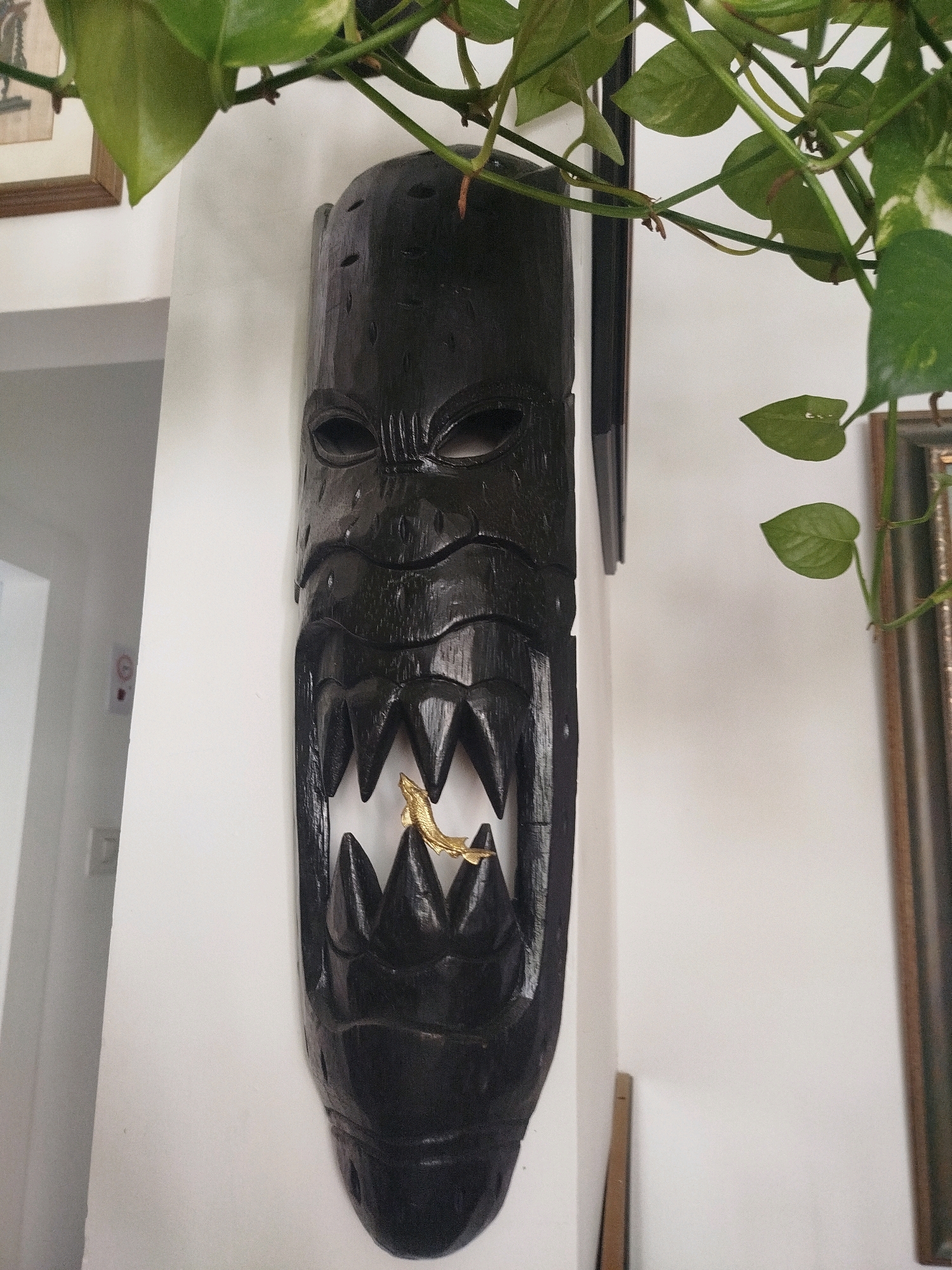 The image shows a close-up of a dark, carved wooden mask with an open mouth, mounted on a white wall. Inside the mask's mouth, a small, golden, fish-shaped object is visible. Green leaves from a plant hang from the top of the frame, partially obscuring the top of the mask and the wall. The background is predominantly white, with a hint of a doorway or opening on the left side and a framed object visible on the right. There are no people or animals present in the image. The environment appears to be indoors, possibly a home or a display area. There is no information in the image to suggest a specific location within the city of Ashkelon, Israel.