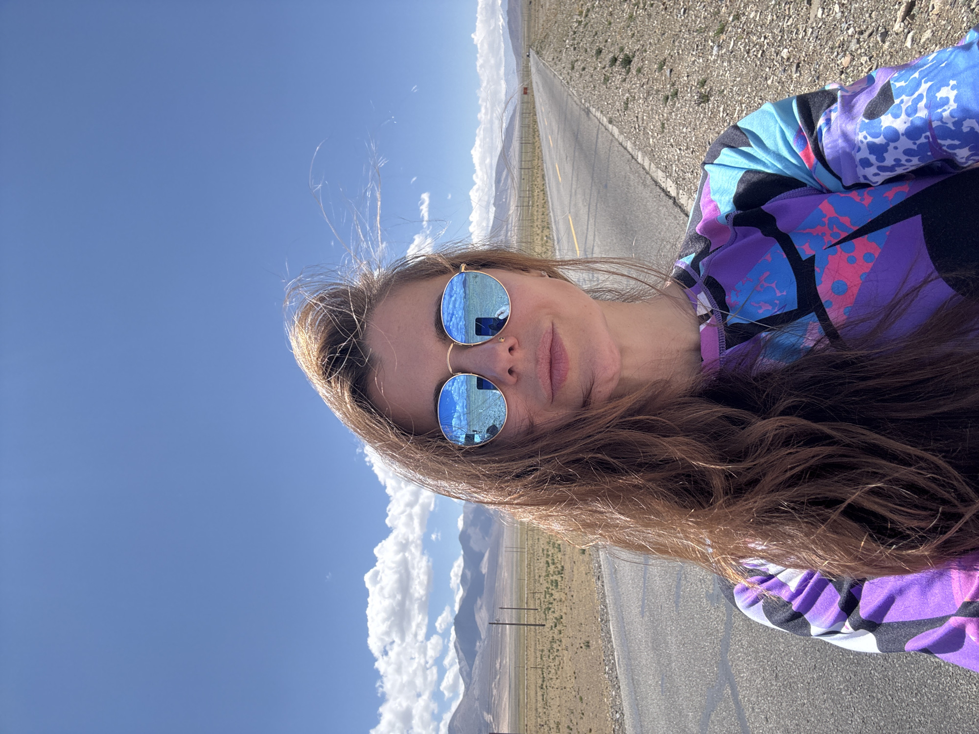 The image, rotated 90 degrees counter-clockwise, is a bright daylight selfie of a person in an open, arid landscape, likely Mainpu township, China. The subject is a female with long, flowing brown hair, partially blowing in the wind. She is looking towards the camera with a neutral expression and light pink lips, and a small nose piercing is visible. She wears round, mirrored sunglasses that reflect the clear blue sky, white clouds, and a distant, mountainous terrain. Her attire consists of a vibrant, multi-colored hoodie featuring an abstract pattern in shades of purple, blue, pink, and black.
The background shows a straight, asphalt road with a yellow dividing line, stretching into the distance towards what appears to be a horizon lined with mountains and sparse vegetation. The terrain on either side of the road is dry and rocky. The weather is clear and sunny, indicated by the strong light and clear sky with scattered white clouds. Several thin utility poles or fence posts are visible along the roadside in the middle distance. There is no visible text in the image. The scene suggests a journey or a stop along a road in a high-altitude or desert-like environment.