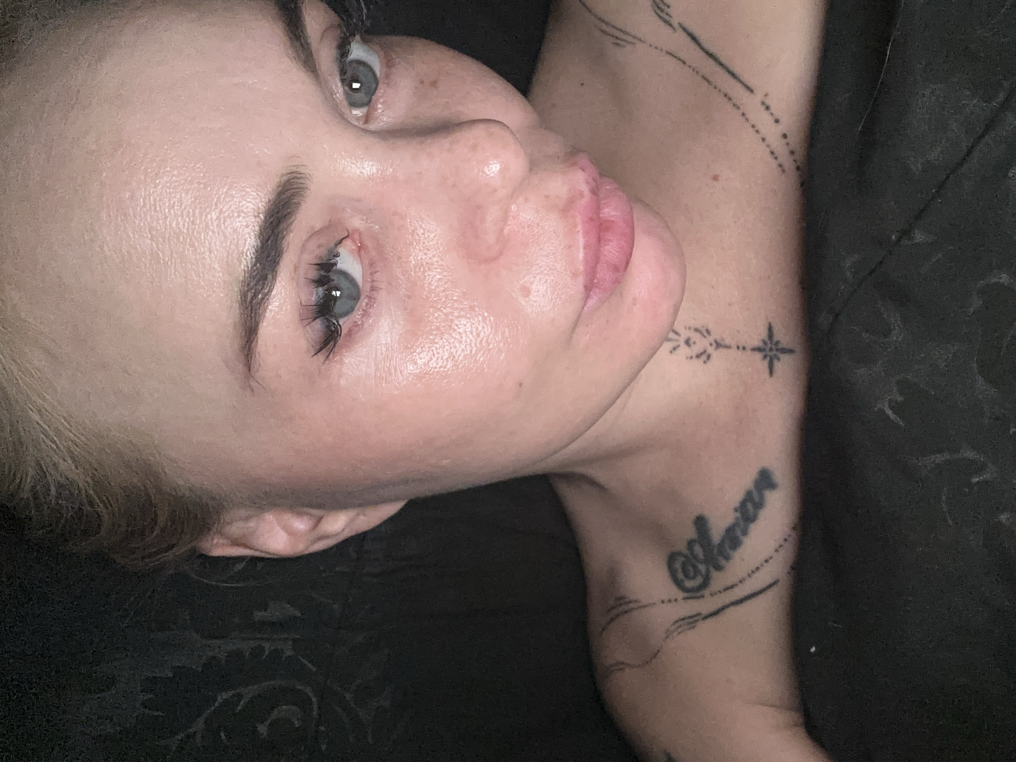 A close-up, overhead, full-face photograph shows a fair-skinned woman with blonde hair pulled back from her face. She has light blue eyes, dark eyebrows, and is wearing false eyelashes. Her skin has a slightly oily sheen and visible freckles. Her lips are a natural pink color. Tattoos are visible on her upper chest and collarbone area. The background is dark and appears to be fabric. The woman is lying down, with her head tilted towards the camera.