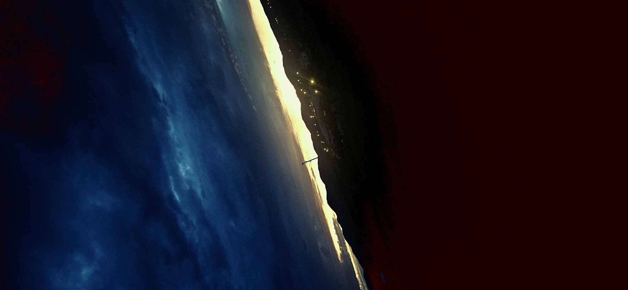 The image captures a dramatic landscape at either dawn or dusk, originally rotated 90 degrees counter-clockwise, presenting the horizon line diagonally. When viewed correctly oriented (rotated 90 degrees clockwise), a vivid horizon stretches across the middle.
The sky, occupying the upper left section, is a deep, rich blue with scattered, textured clouds in lighter blue and purple hues. Along the horizon, a brilliant strip of golden-yellow light glows intensely, indicative of the sun's low position, suggesting either sunrise or sunset.
Below this luminous horizon, on the right, a dark, silhouetted form of hills or mountains is visible. Numerous small, bright yellow lights dot this dark landmass, suggesting human settlements or structures in the distance. To the left of the bright horizon, a vast expanse of dark water or a reflective surface mirrors the deep blue tones of the sky. A slender, dark, pole-like structure stands vertically, extending from this dark body of water towards the bright horizon.
The bottom right portion of the image, below the illuminated hills, is a very dark, almost black and deep reddish-brown area, likely representing shadowed foreground terrain or deep perspective.
The scene evokes a serene and atmospheric mood, characterized by the strong contrast between light and shadow and the vibrant blues, purples, and golden-yellows. There are no people visible, nor any discernible text. Given the location context of Momin Prohod, Bulgaria, an inland town, the body of water is likely a large lake, reservoir, or a river valley reflecting the sky, with mountains forming the silhouette.